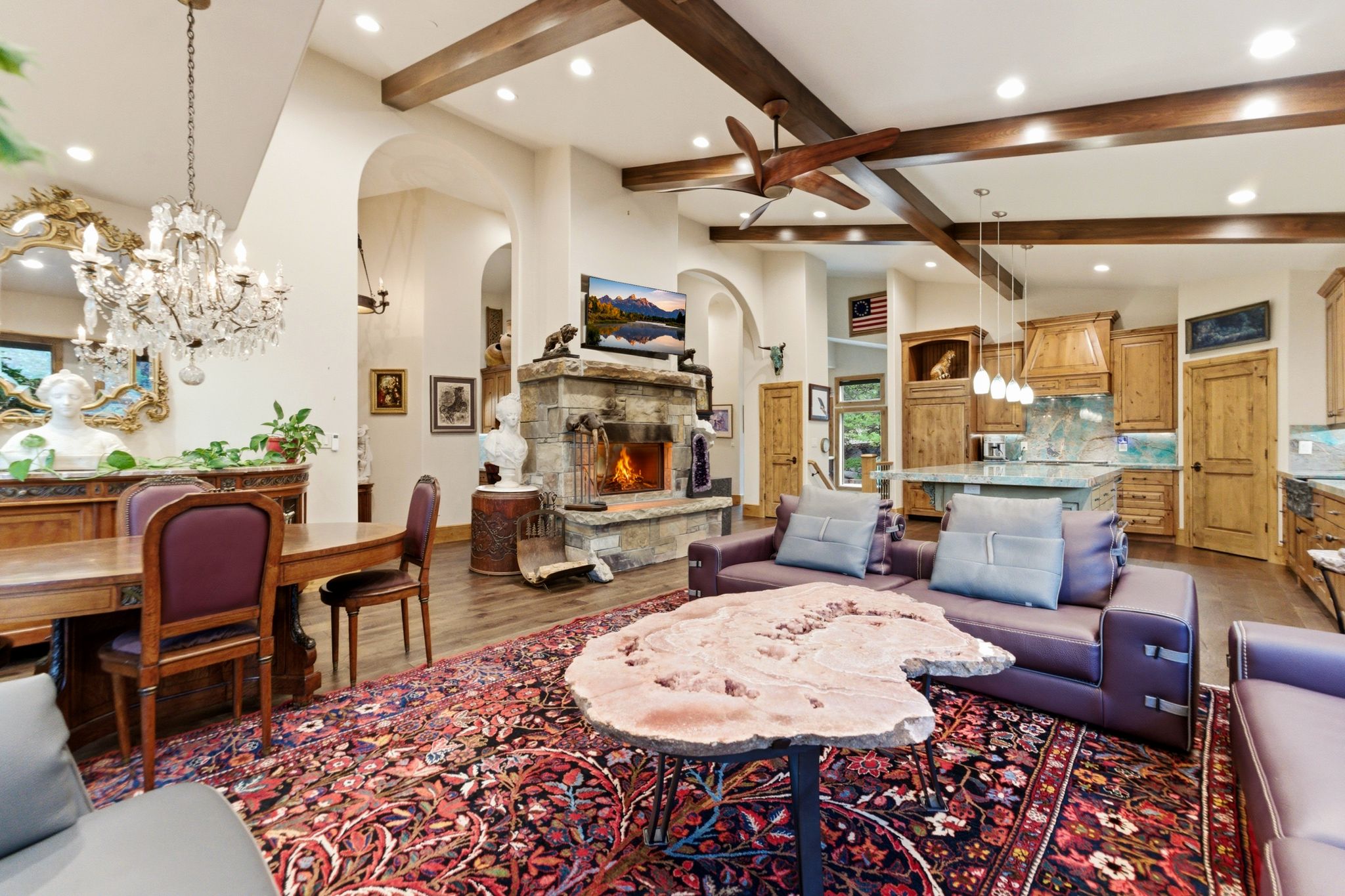 Vaulted ceilings with exposed wood beams, a stone fireplace with a wood mantle, and wood-finish flooring define this open-concept living area