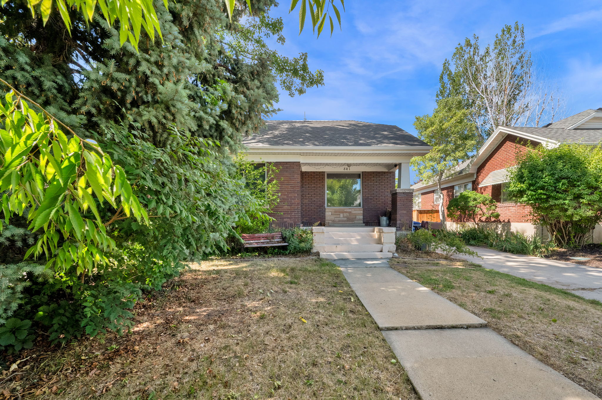 Brick facade residence featuring a covered front porch, concrete walkway, established front yard landscaping, and a pitched roof