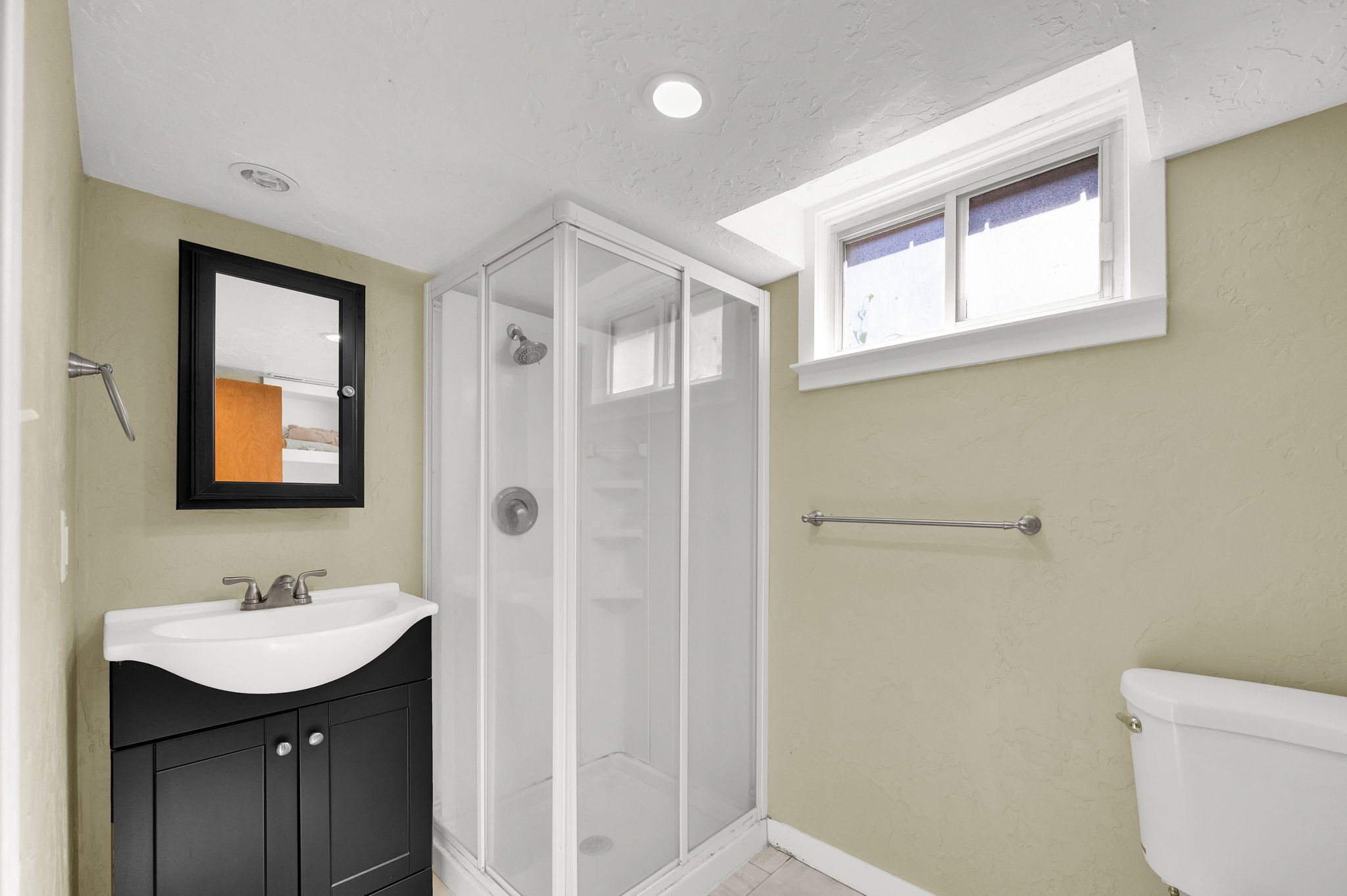Modern bathroom featuring a compact glass-enclosed shower, a vanity with a dark wood-finish cabinet and integrated sink, a framed medicine cabinet, a functional window, and recessed lighting