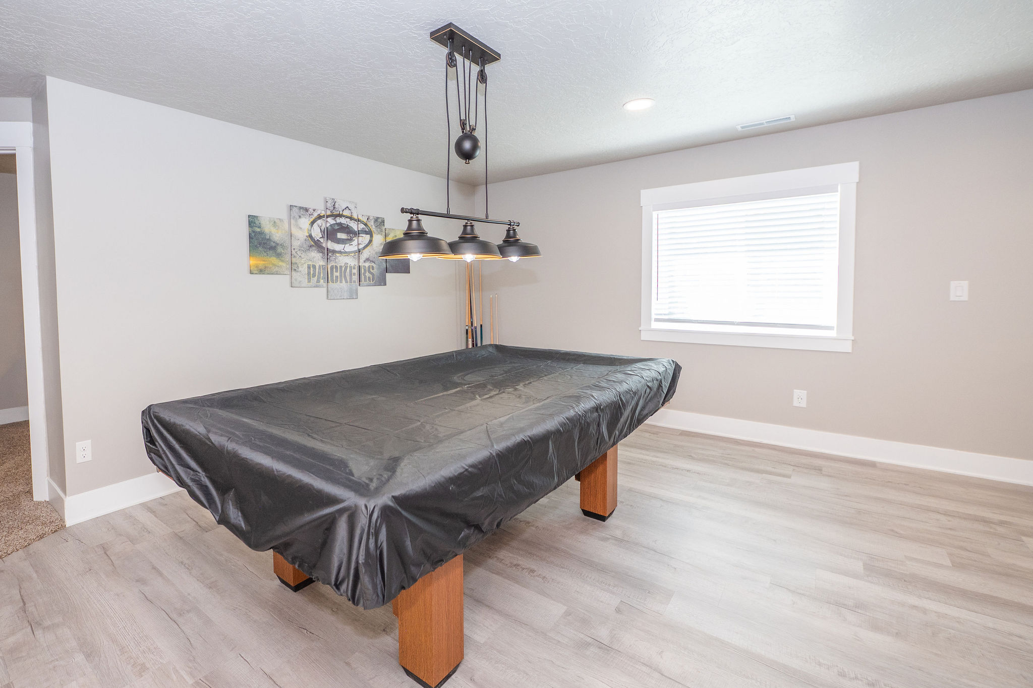 Spacious recreation area featuring light wood-finish flooring, a contemporary industrial-style light fixture, and a window with blinds