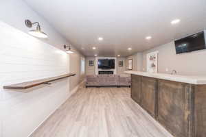 Open-concept lower level featuring shiplap accent wall with sconce lighting, wood-finish flooring, a built-in wet bar with sink, and recessed lighting