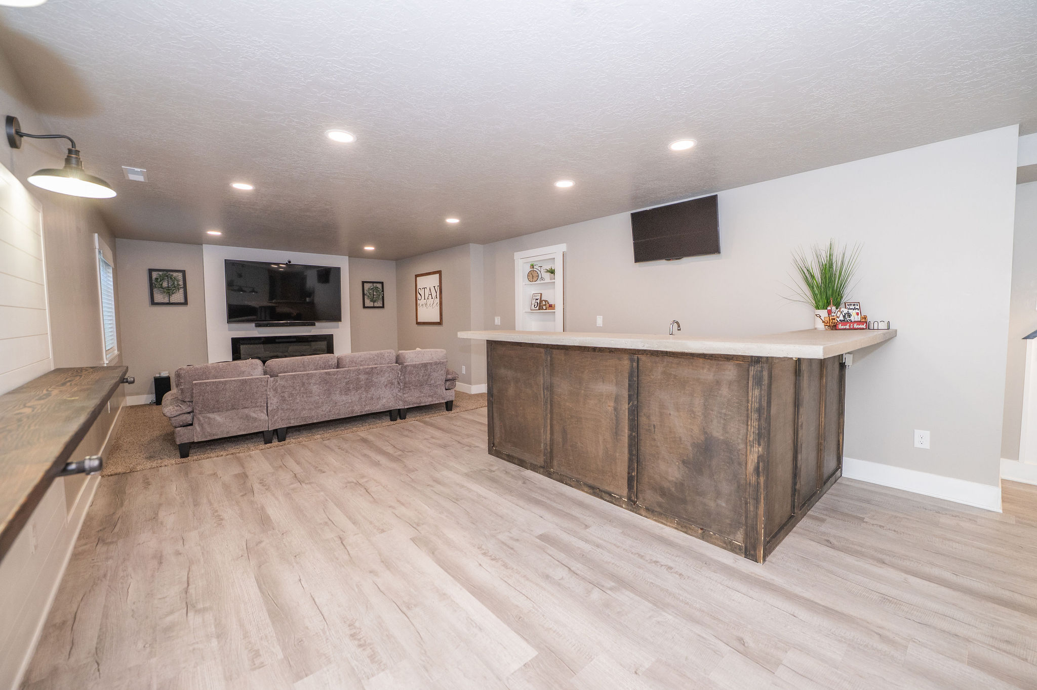 Expansive entertainment space featuring a custom bar with a light-toned countertop, a built-in electric fireplace with a white mantel, recessed lighting, and wood-finish flooring