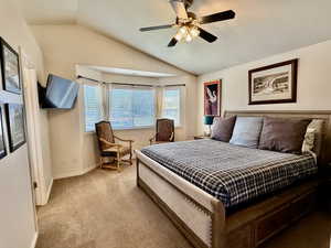 Vaulted ceiling bedroom featuring a bay window with blinds, a ceiling fan with integrated lighting, a wall-mounted television, and carpet flooring