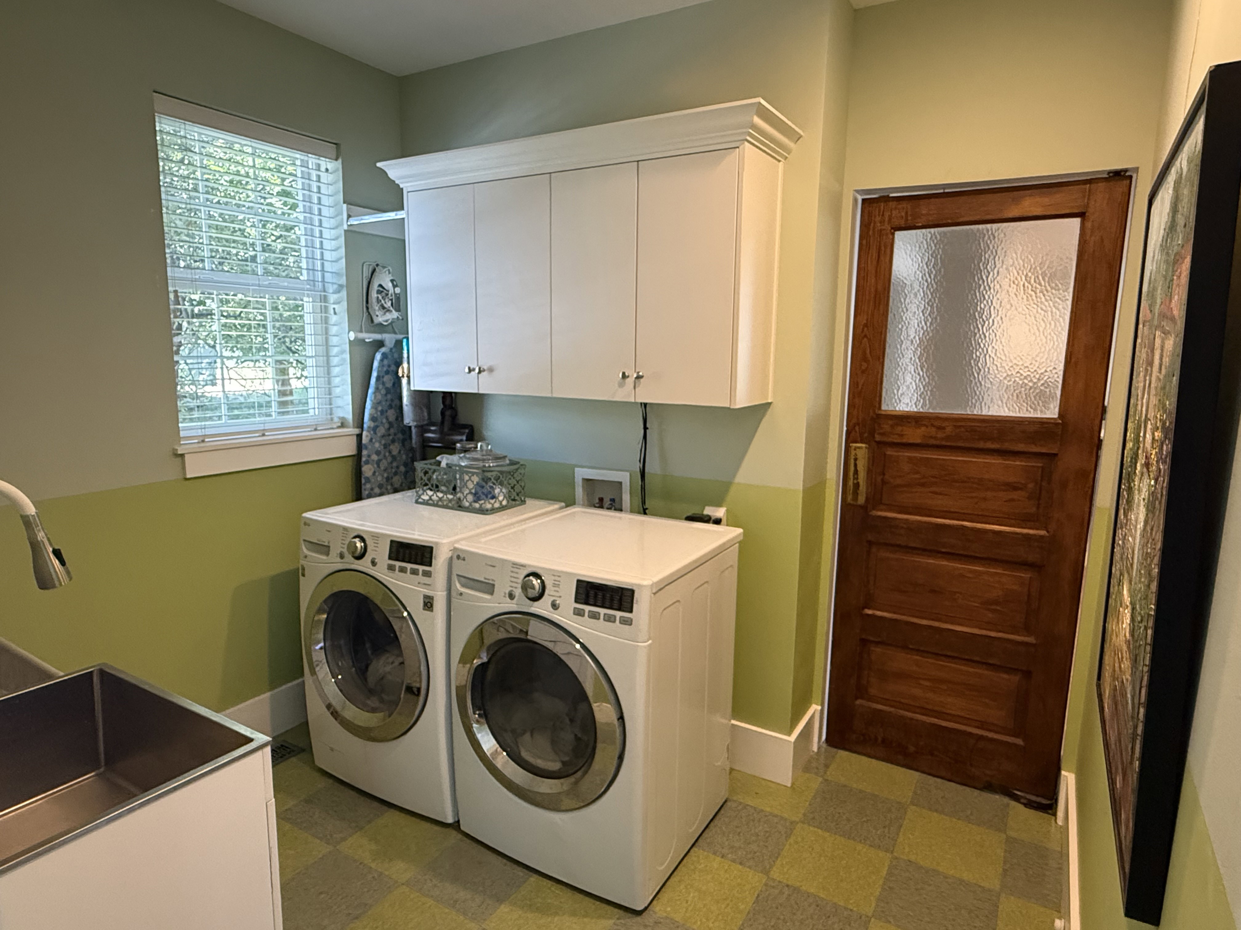 Dedicated laundry room featuring checkered flooring, a window with horizontal blinds, overhead white cabinetry, and a utility sink with a pull-down faucet