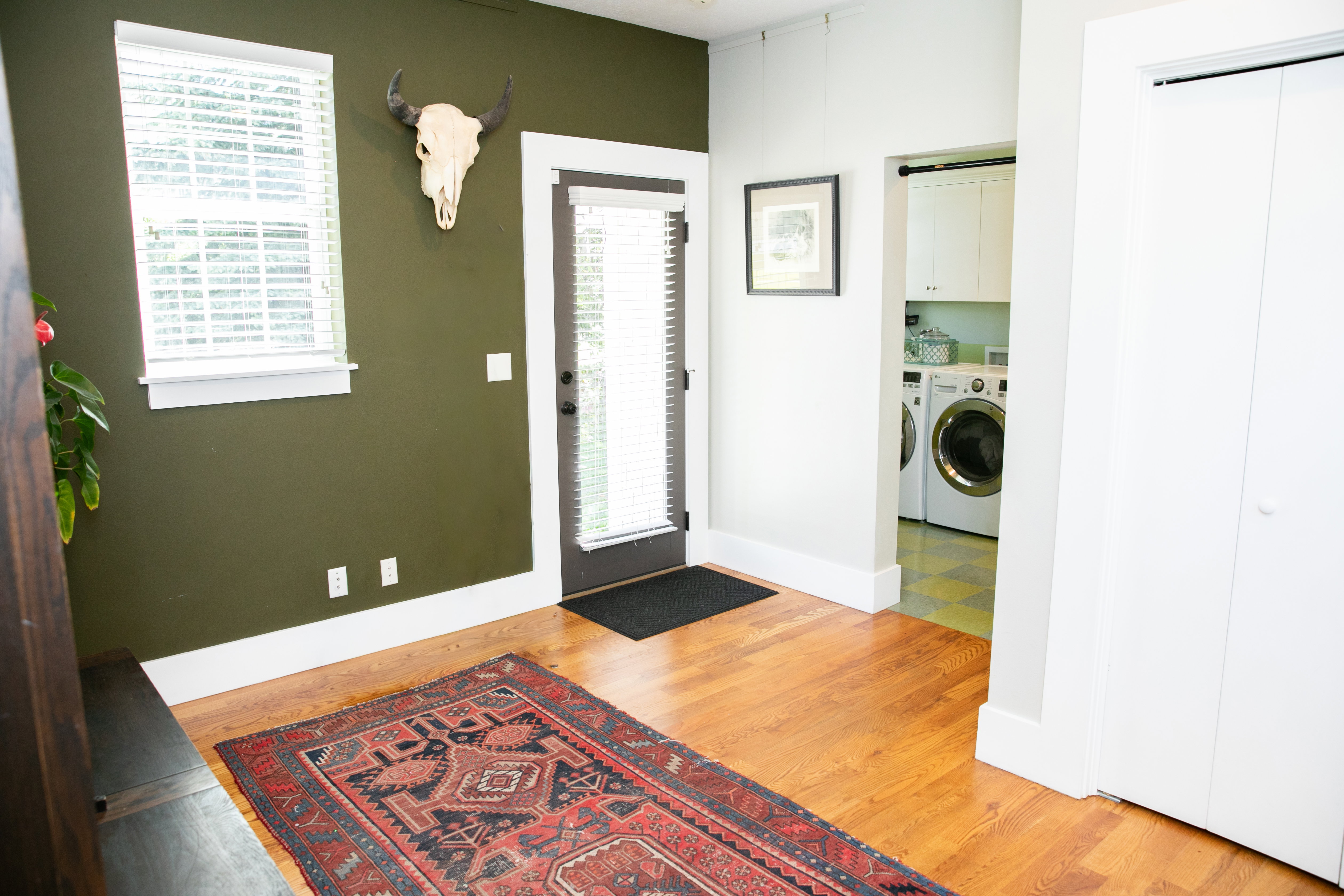 Rich hardwood flooring throughout, featuring a window with blinds, a dark-framed exterior door, and a dedicated laundry area with built-in cabinetry and tile flooring