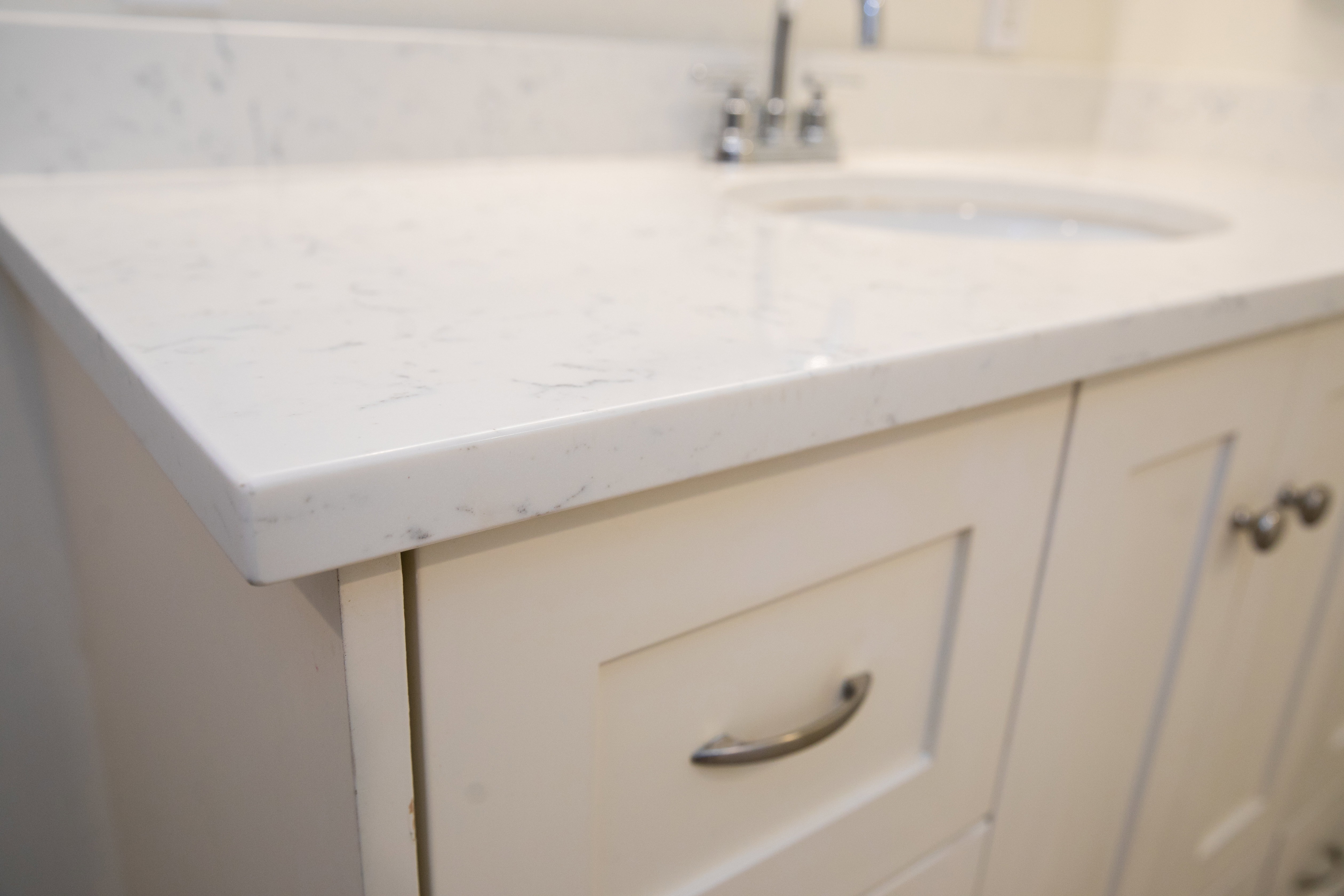 White countertop with subtle veining, undermount sink, chrome faucet, and off-white shaker-style cabinetry with brushed nickel hardware