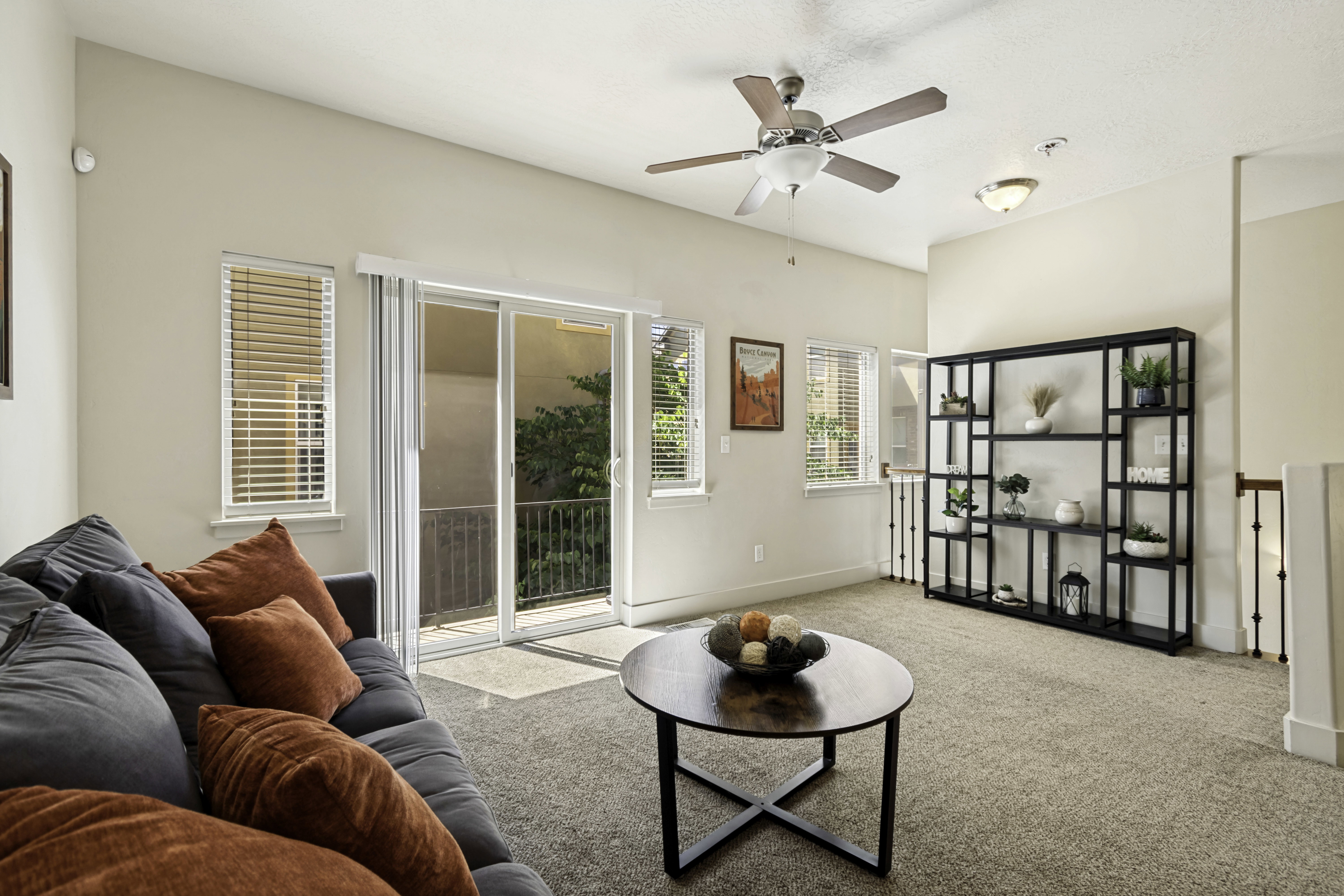 Living area featuring neutral wall tones, textured carpeting, a sliding glass door leading to a balcony, multiple windows with blinds, and a permanent ceiling fan with integrated lighting
