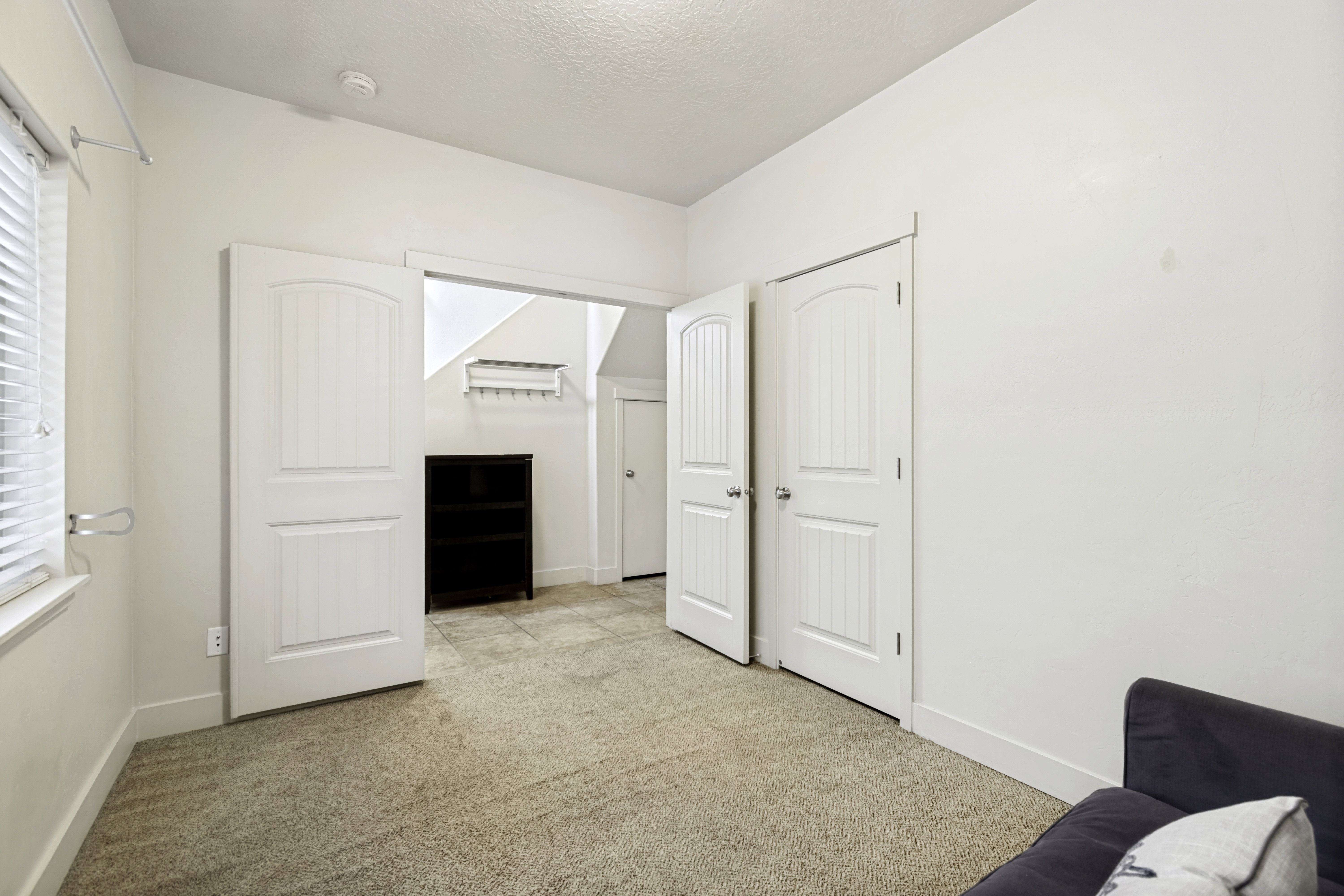 Carpeted room with white walls, featuring a window with blinds, two white paneled doors, and an open doorway leading to a tiled area with a dark built-in shelving unit