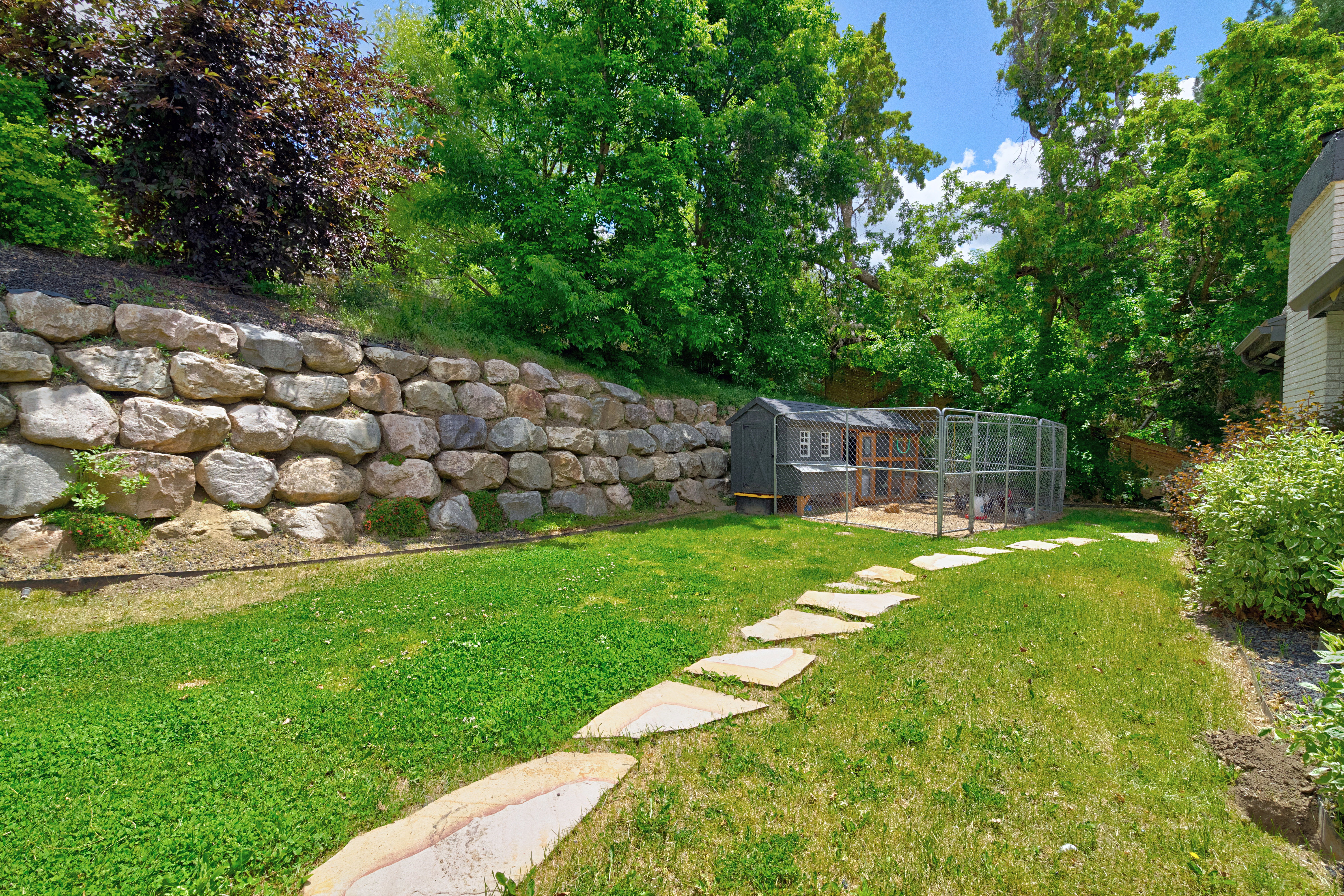 Expansive backyard featuring a stone retaining wall, mature trees, a stepping stone path, and a functional animal enclosure