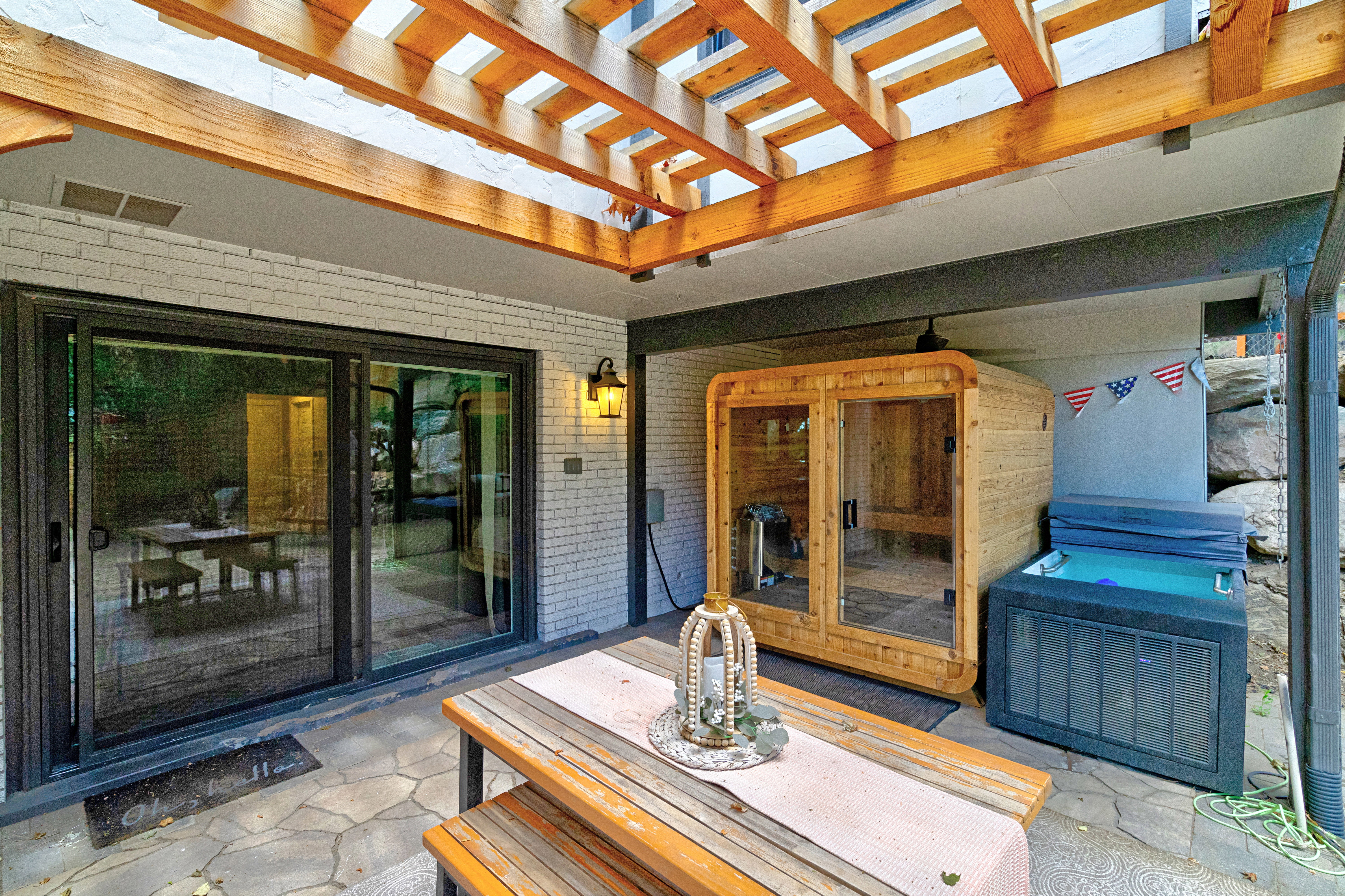 Covered patio area featuring a slatted wood pergola, stone-paver flooring, and white painted brick