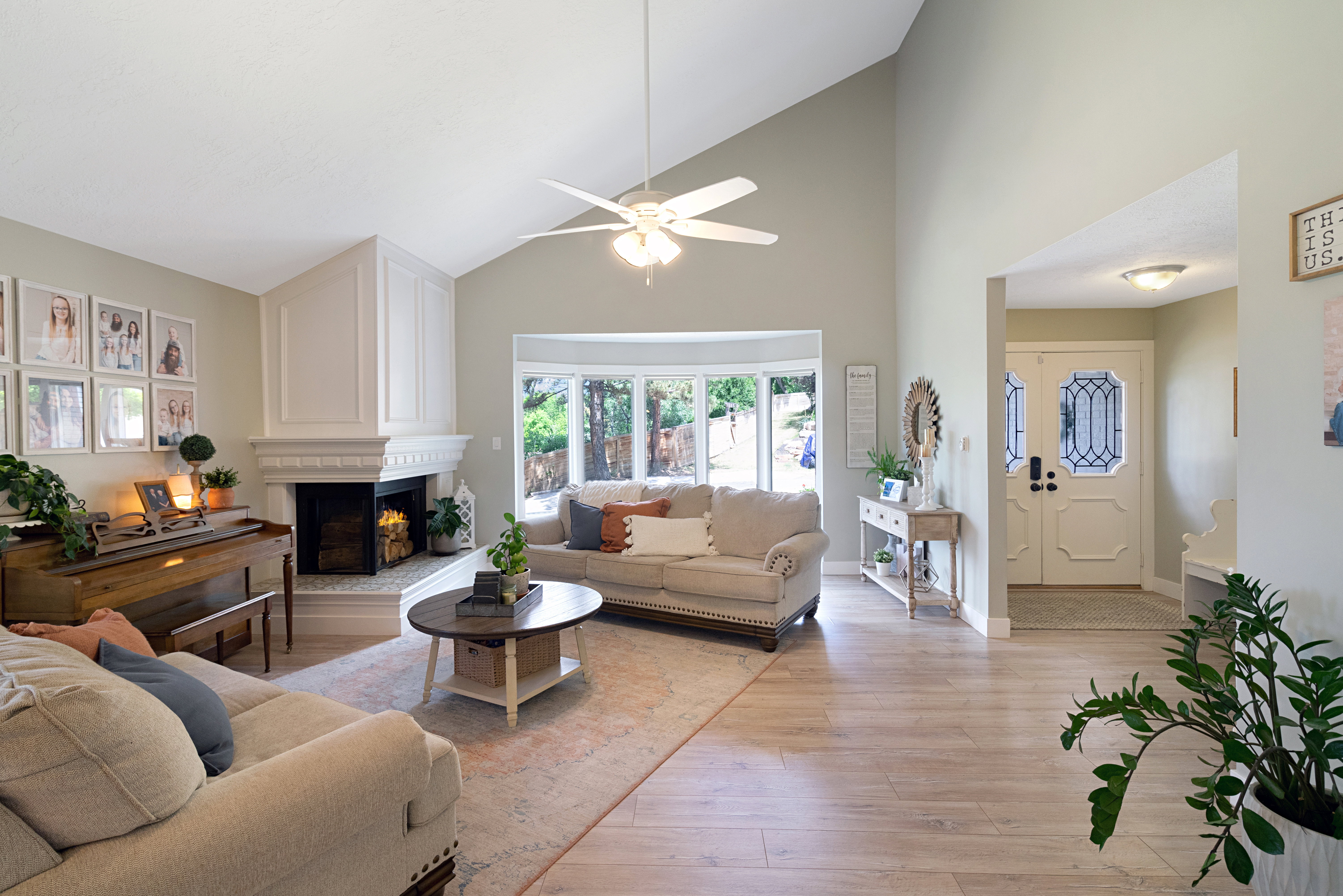 Living area featuring vaulted ceilings, wood-finish flooring, a decorative fireplace with a mantel, a bay window, and a ceiling fan