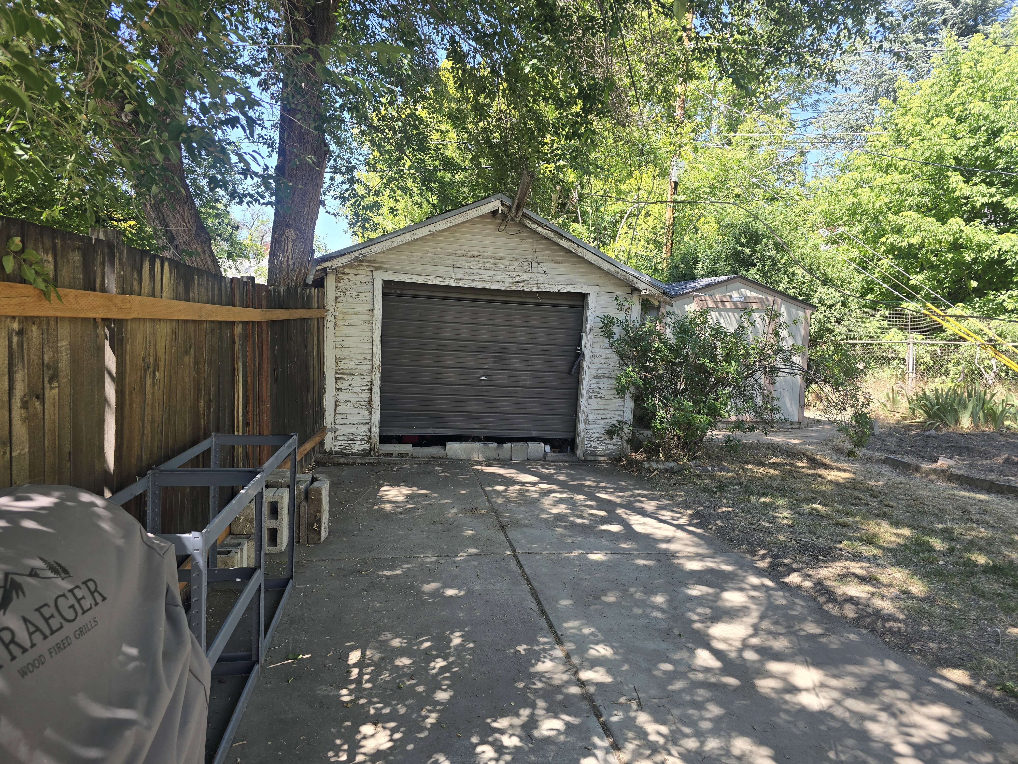 Detached garage with roll-up door, adjacent storage shed, concrete driveway, wood privacy fence, and mature trees