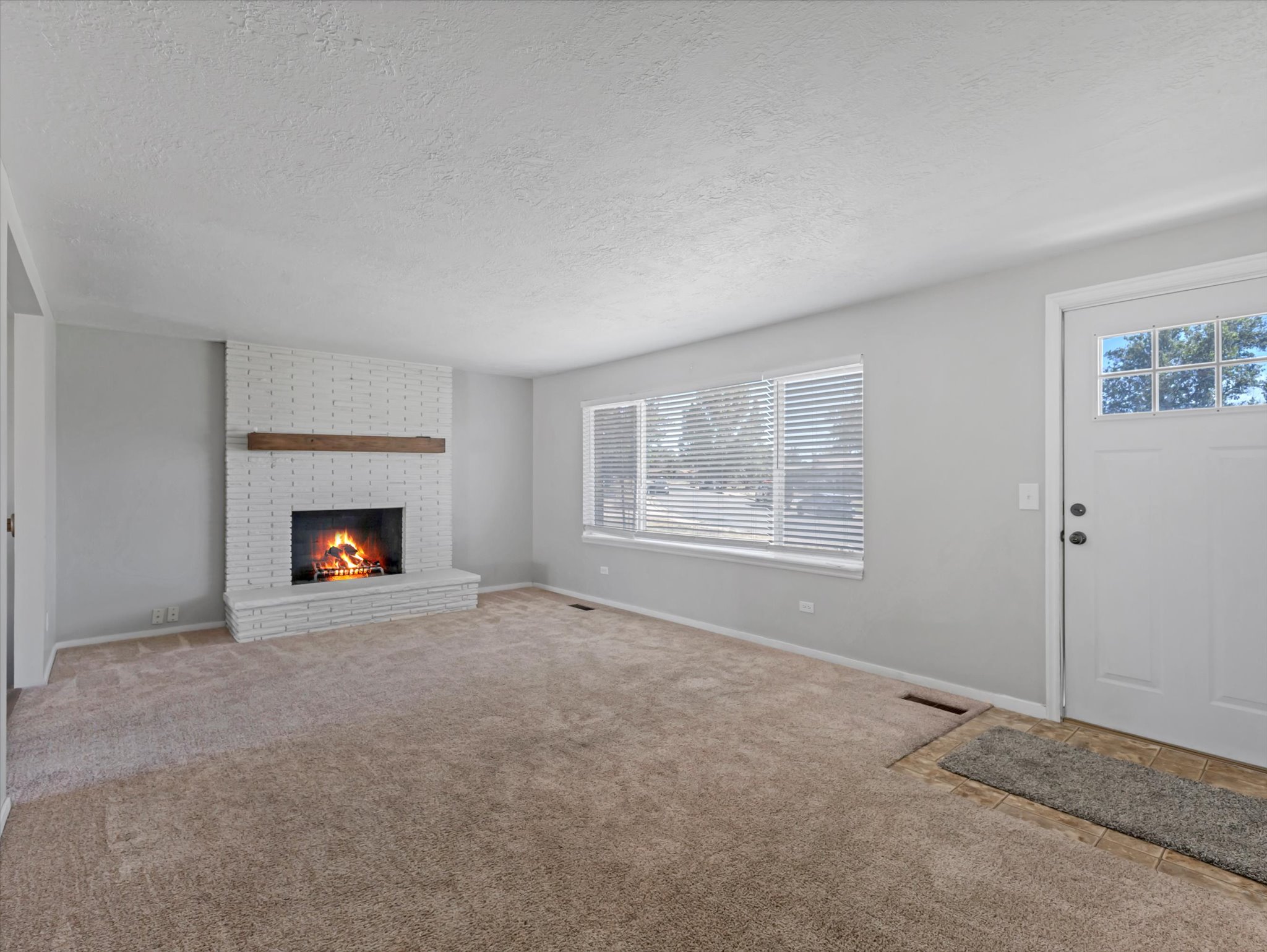 Living area featuring a white brick fireplace with a wood mantel, carpeted flooring, a multi-pane front door, and a large window with horizontal blinds