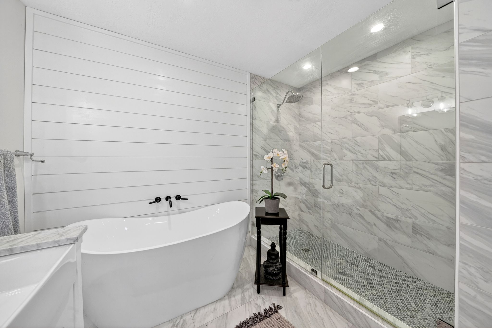 Contemporary bathroom featuring a freestanding soaking tub with wall-mounted matte black fixtures, a shiplap accent wall, and a spacious glass-enclosed shower with large format marble-look tile walls and a mosaic pebble floor