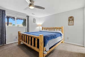 Bedroom featuring a window with a dark curtain, light gray walls, a ceiling fan with integrated lighting, and neutral-toned carpeting