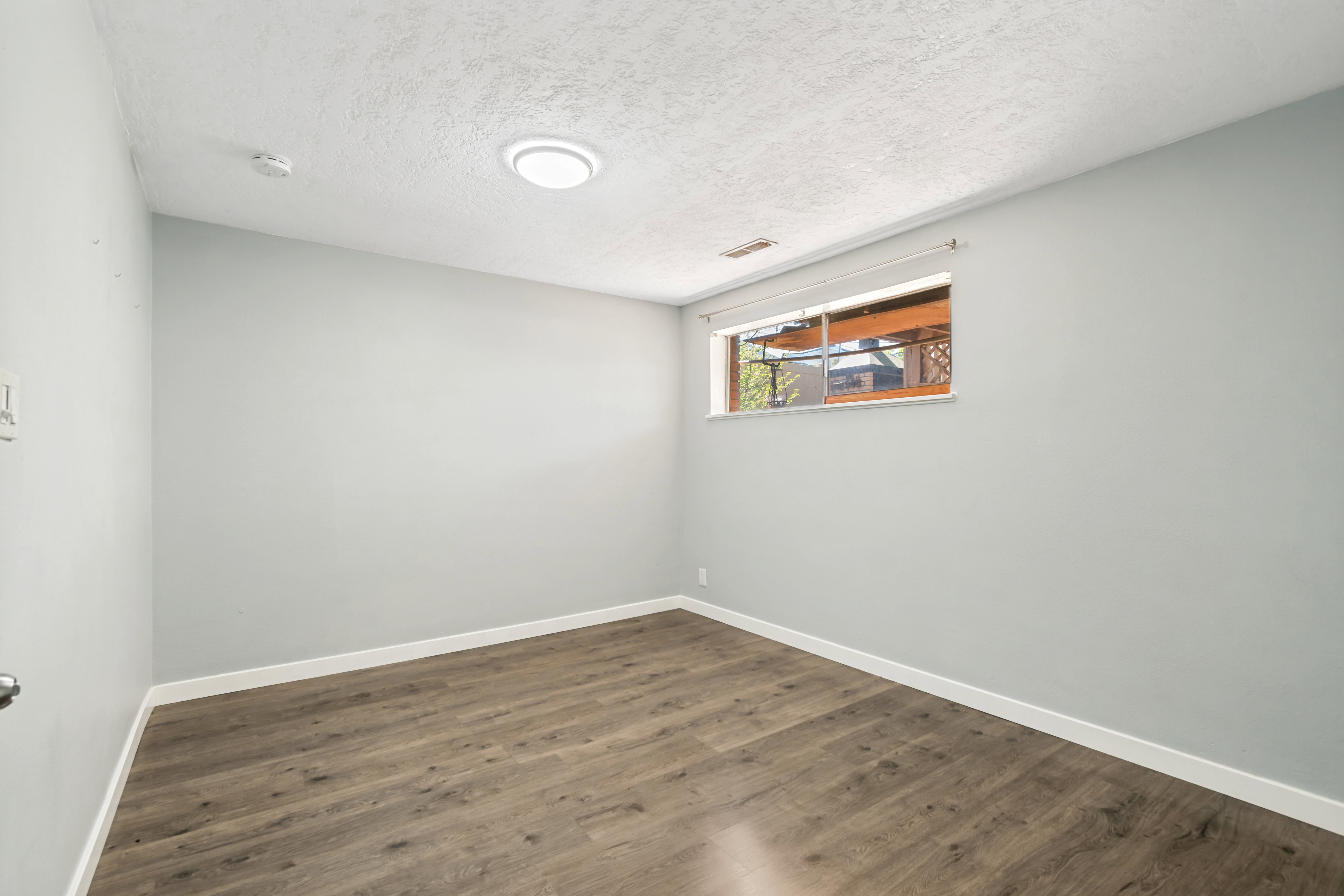 Spacious room featuring wood-finish flooring, light grey painted walls, white baseboards, and a window with natural light