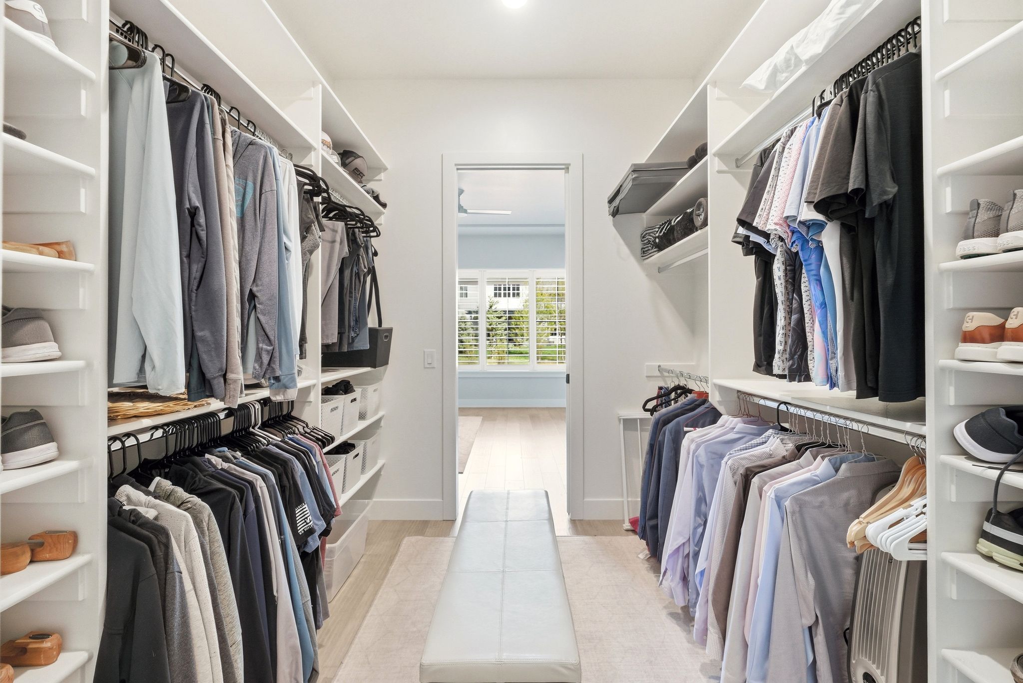 Spacious walk-in closet featuring extensive built-in shelving, multiple hanging rods for garment organization, integrated shoe racks, a central bench, and light wood-finish flooring