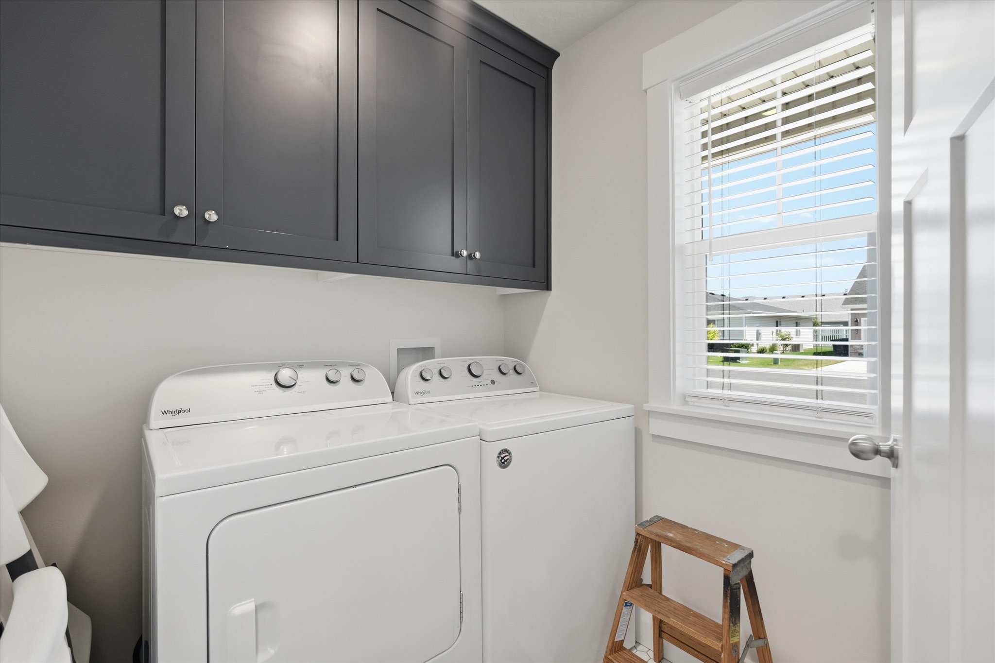 Laundry area featuring dark gray upper cabinetry with silver-tone hardware, white walls, and a white framed window with horizontal blinds