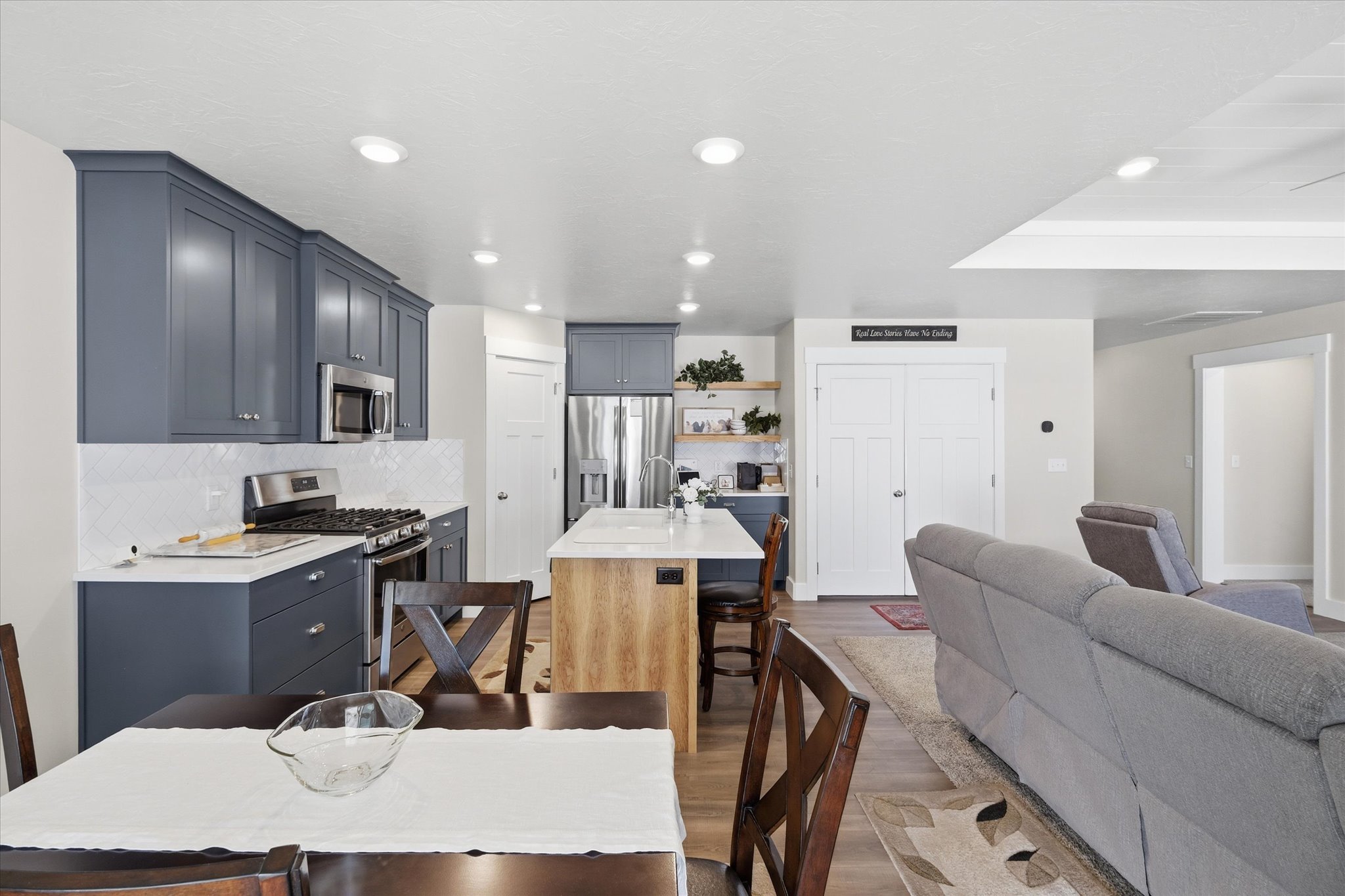 Open-concept kitchen and dining area featuring dark blue cabinetry, stainless steel appliances, a white tile backsplash, a central island with an integrated breakfast bar, and wood-finish flooring