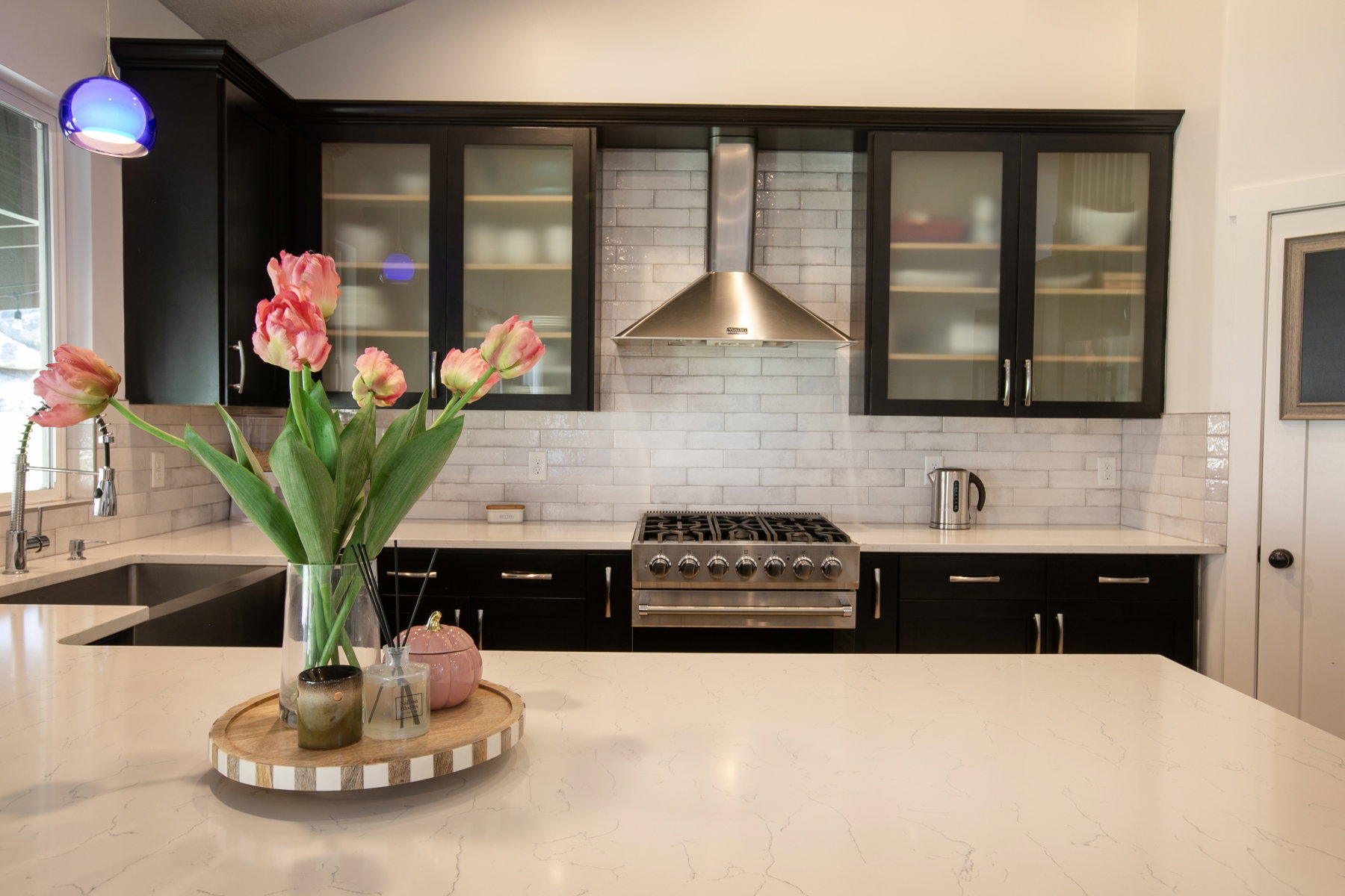 Kitchen featuring dark cabinetry with glass-front upper cabinets, white subway tile backsplash, and a stainless steel range with matching hood