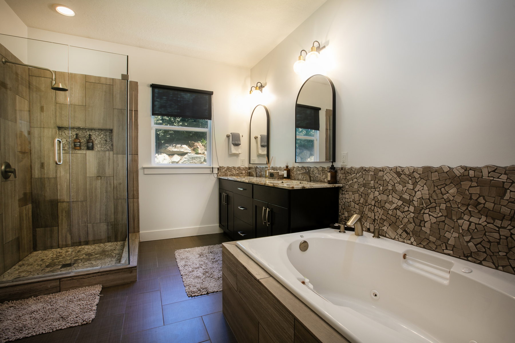 Spacious bathroom featuring a glass-enclosed shower with wood-finish tile, a built-in jetted tub, a dual vanity with granite countertops, and an accent wall with mosaic tile