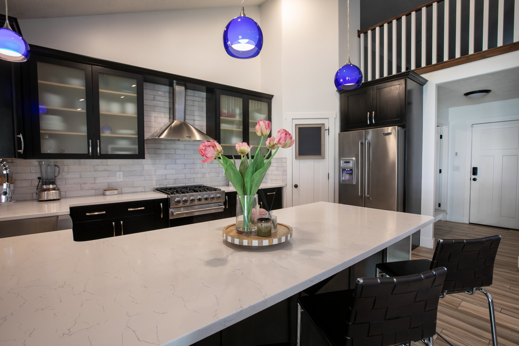 Kitchen featuring a spacious island with a light-toned countertop, dark cabinetry with glass upper doors, a stainless steel range with a ventilation hood, and white subway tile backsplash