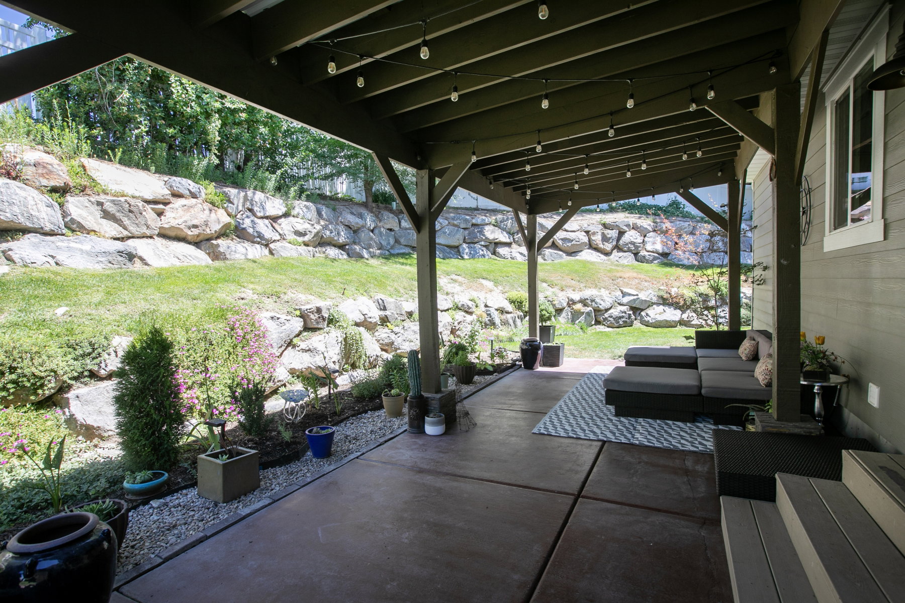 Covered patio featuring string lighting, concrete flooring, and an adjacent rock wall landscaping