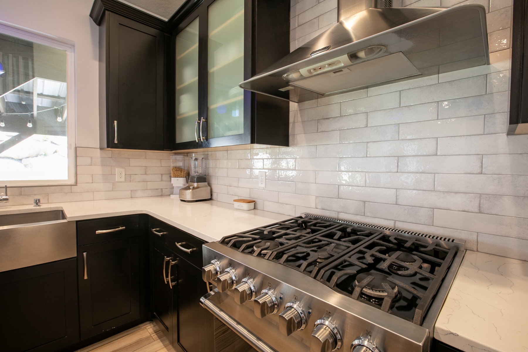 Stainless steel range with a professional-grade hood, dark cabinetry with glass-front uppers, light countertops, subway tile backsplash, and a large stainless steel farmhouse sink