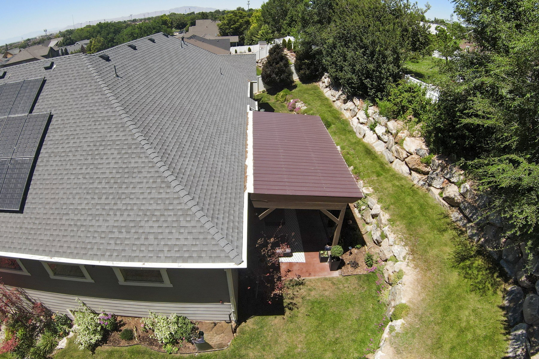 Exterior showcasing a gray shingle roof with integrated solar panels, a brown-roofed covered patio, a rock retaining wall, and established landscaping