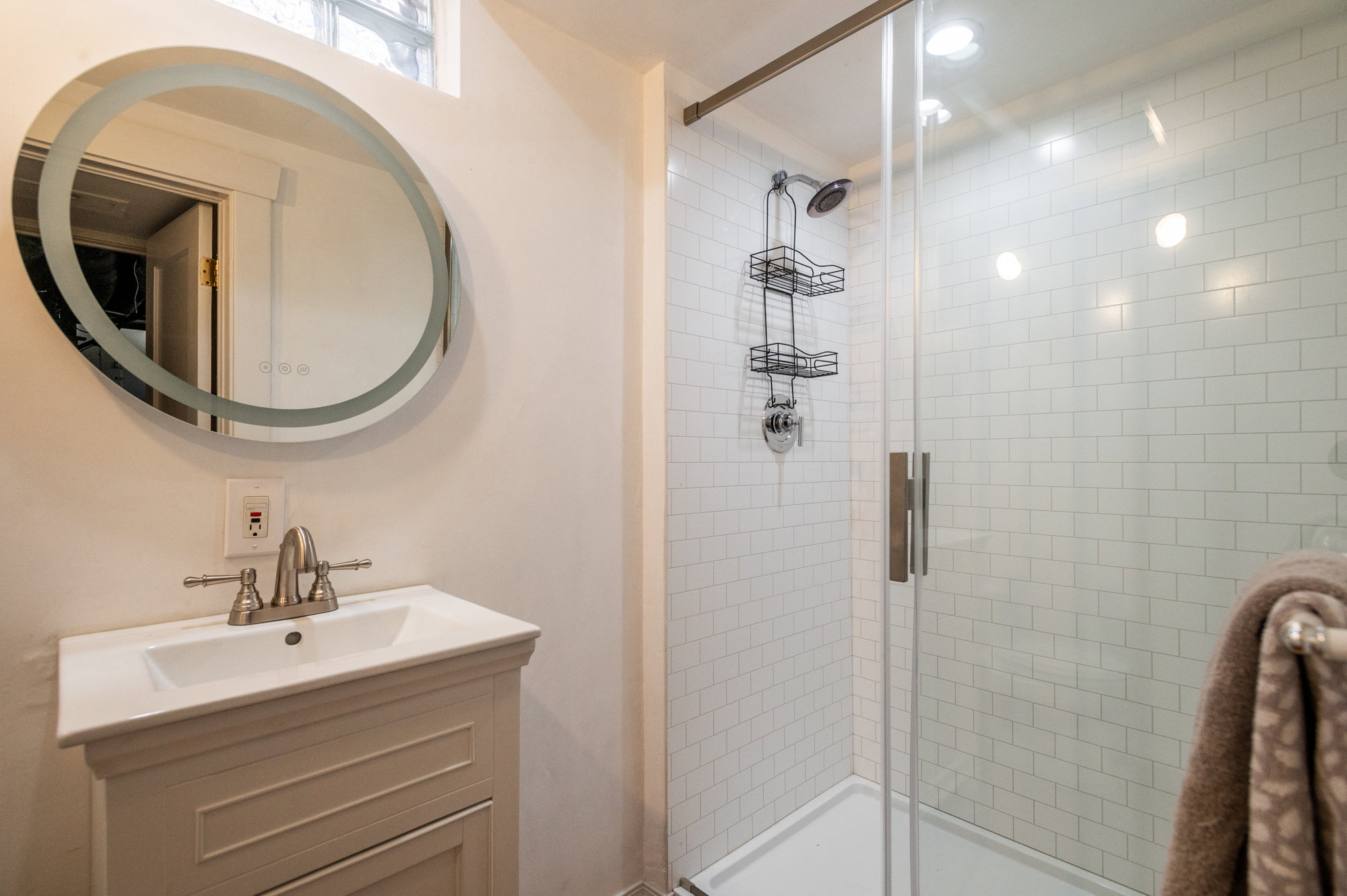 Modern bathroom featuring a glass-enclosed shower with white subway tile, a vanity with a white integrated sink, a brushed nickel faucet, and an illuminated oval mirror