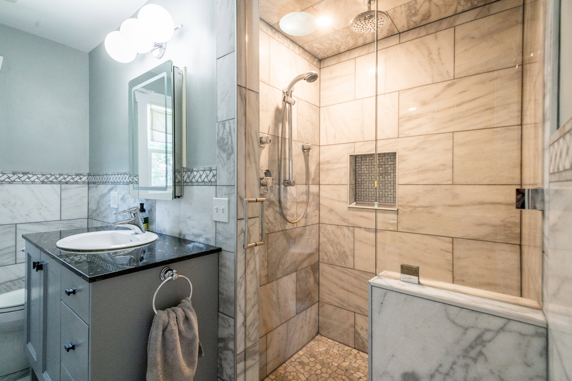 Walk-in shower featuring a rainfall showerhead, handheld sprayer, tiled niche, and stone pebble flooring