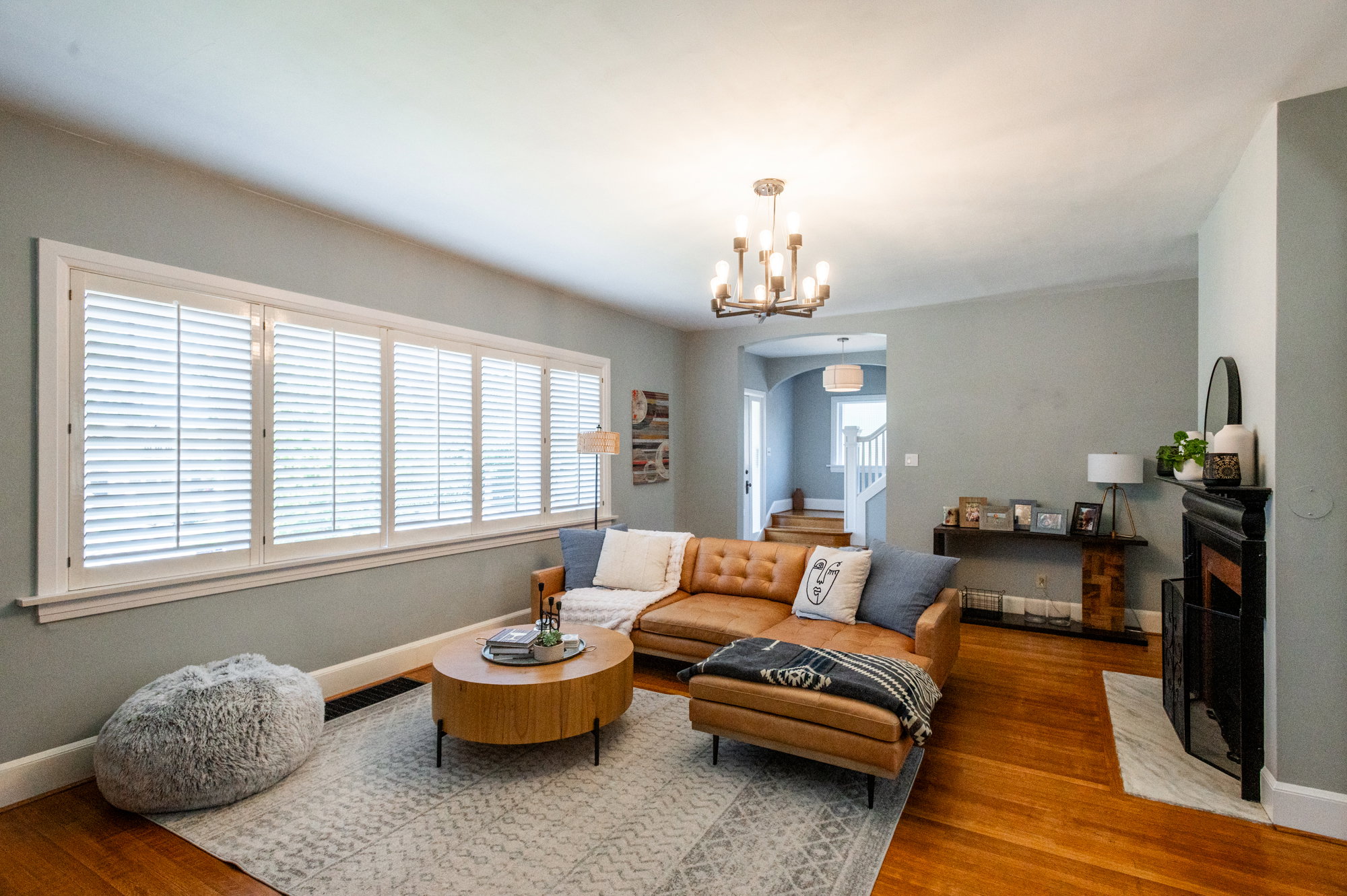 Living area featuring wide plank wood-finish flooring, a white window frame with louvered shutters, a black fireplace with a white hearth, and a multi-arm chandelier