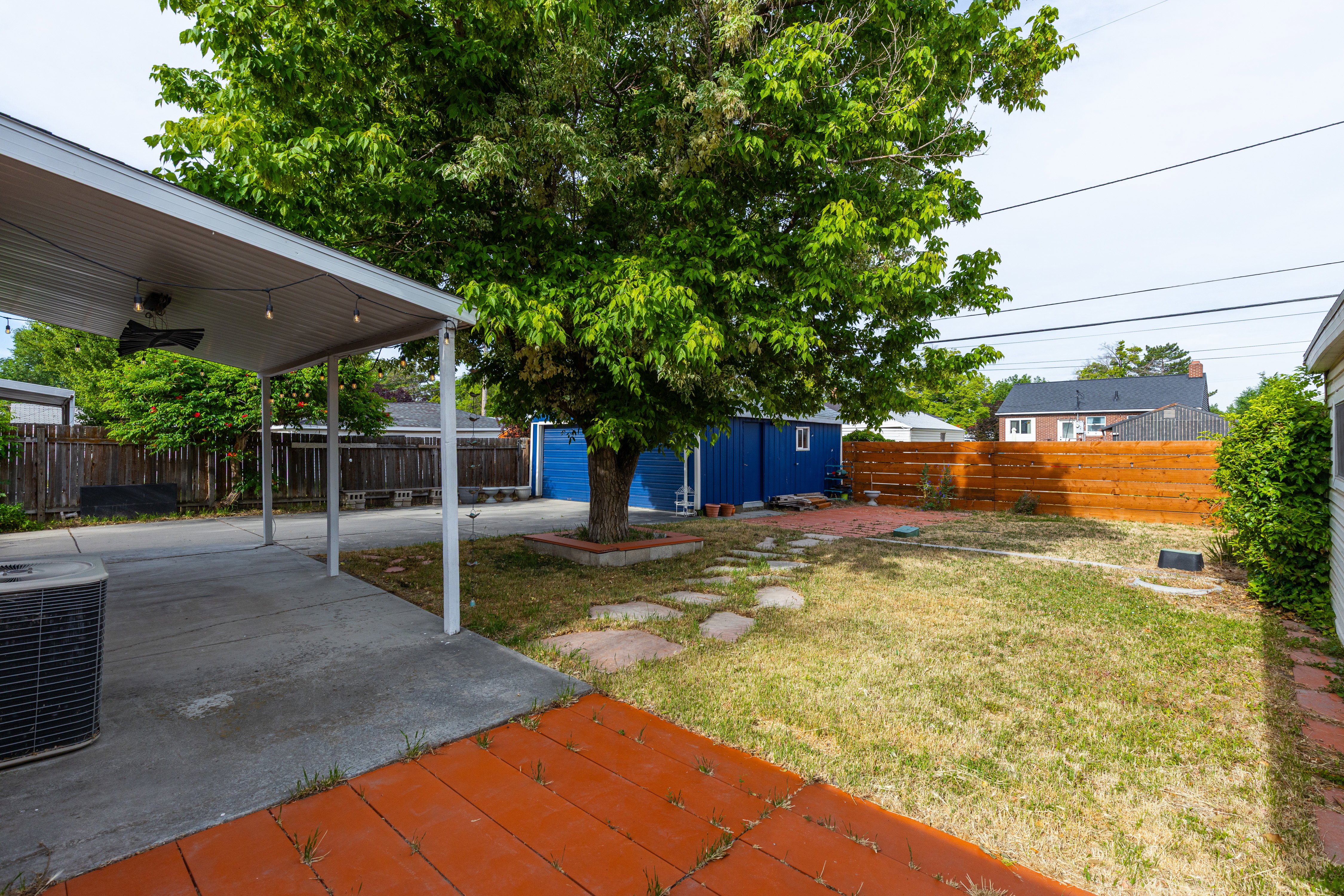 Expansive backyard featuring a mature shade tree, a covered concrete patio, a blue detached structure, and a wood fence