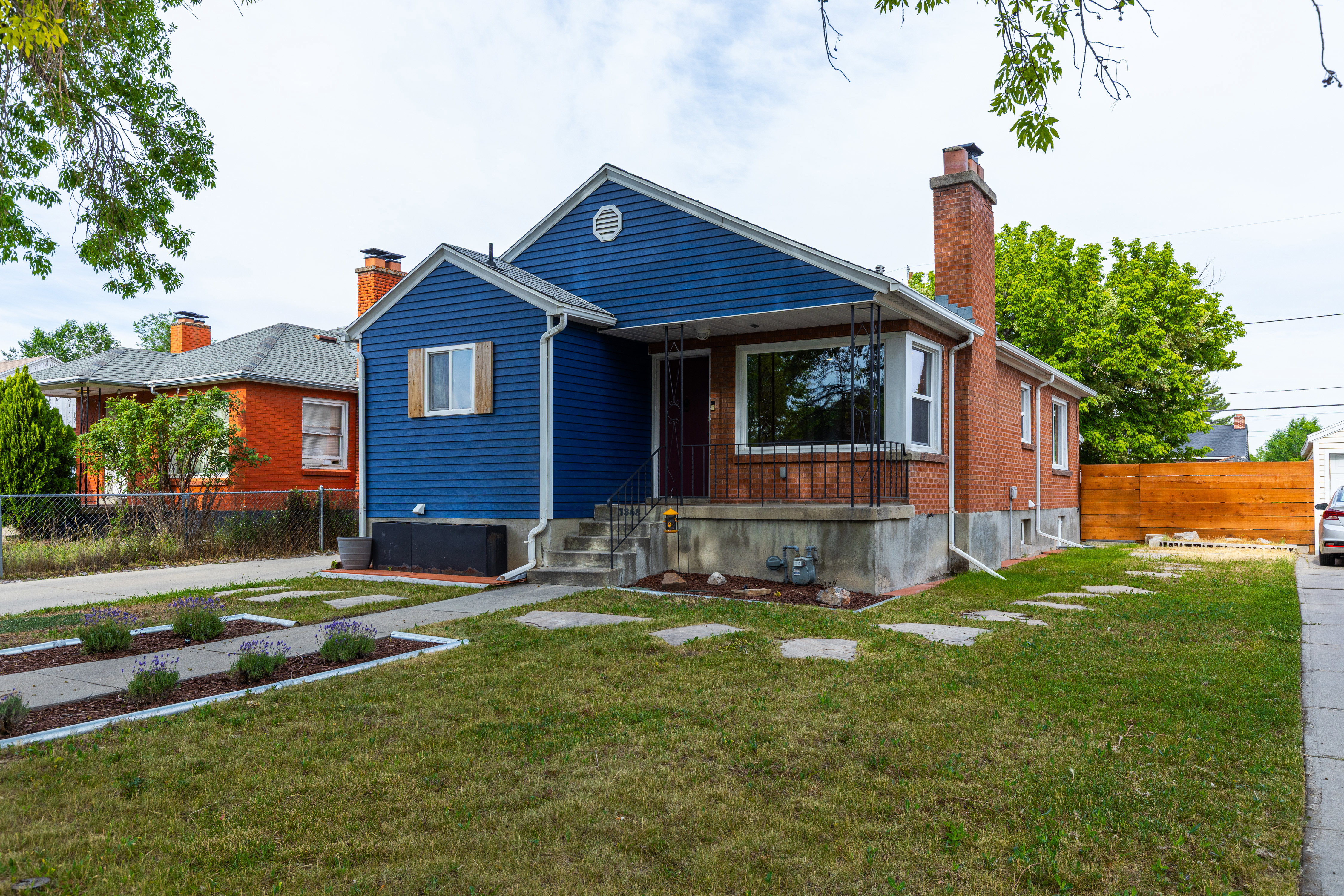 Exterior featuring a striking blue facade with contrasting red brick accents, an inviting front porch with wrought iron railings, a manicured lawn with stepping stones, a concrete driveway, and a newly installed privacy fence