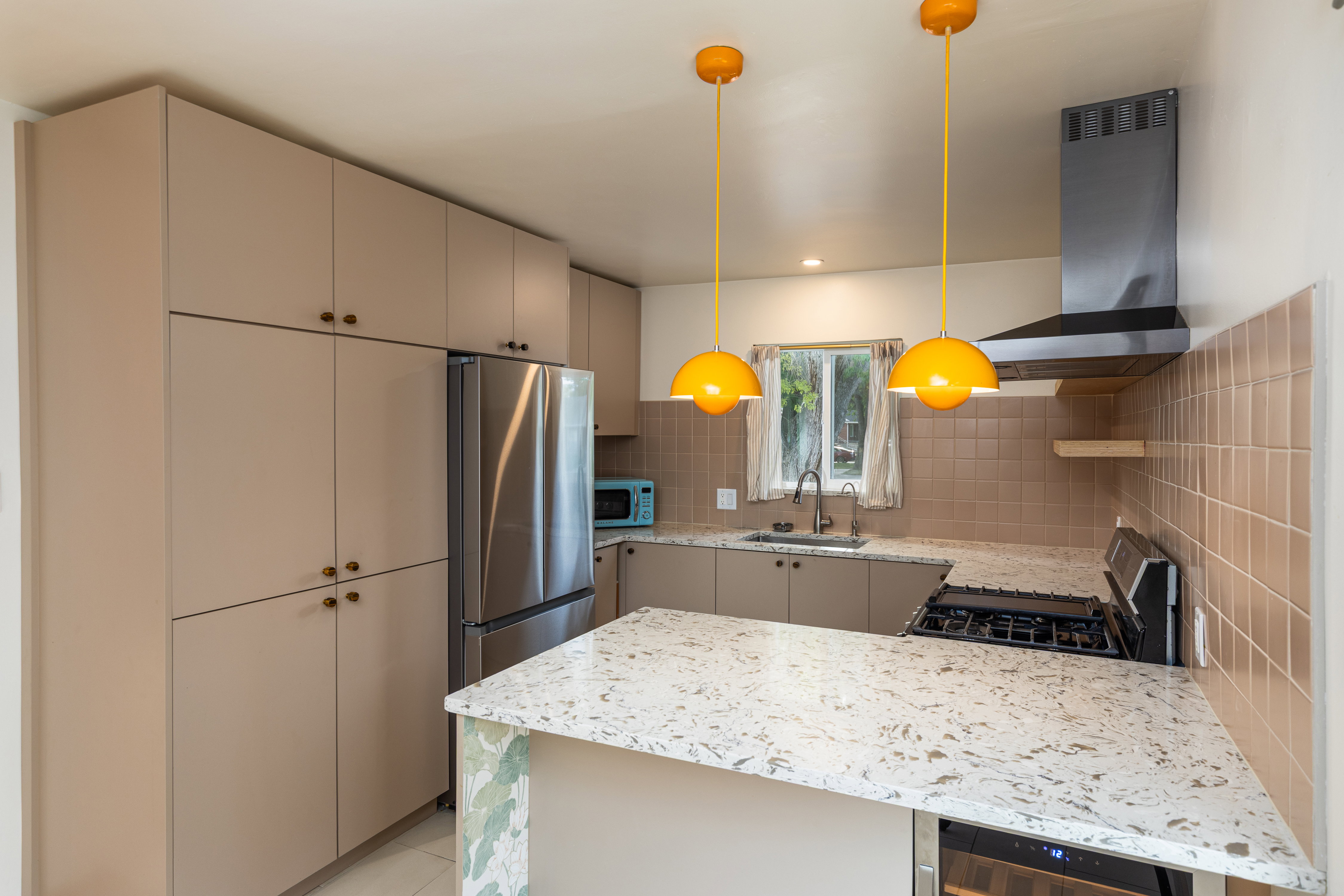 Modern kitchen featuring extensive neutral-toned cabinetry, stainless steel appliances, speckled countertops, a tile backsplash, and a built-in range hood