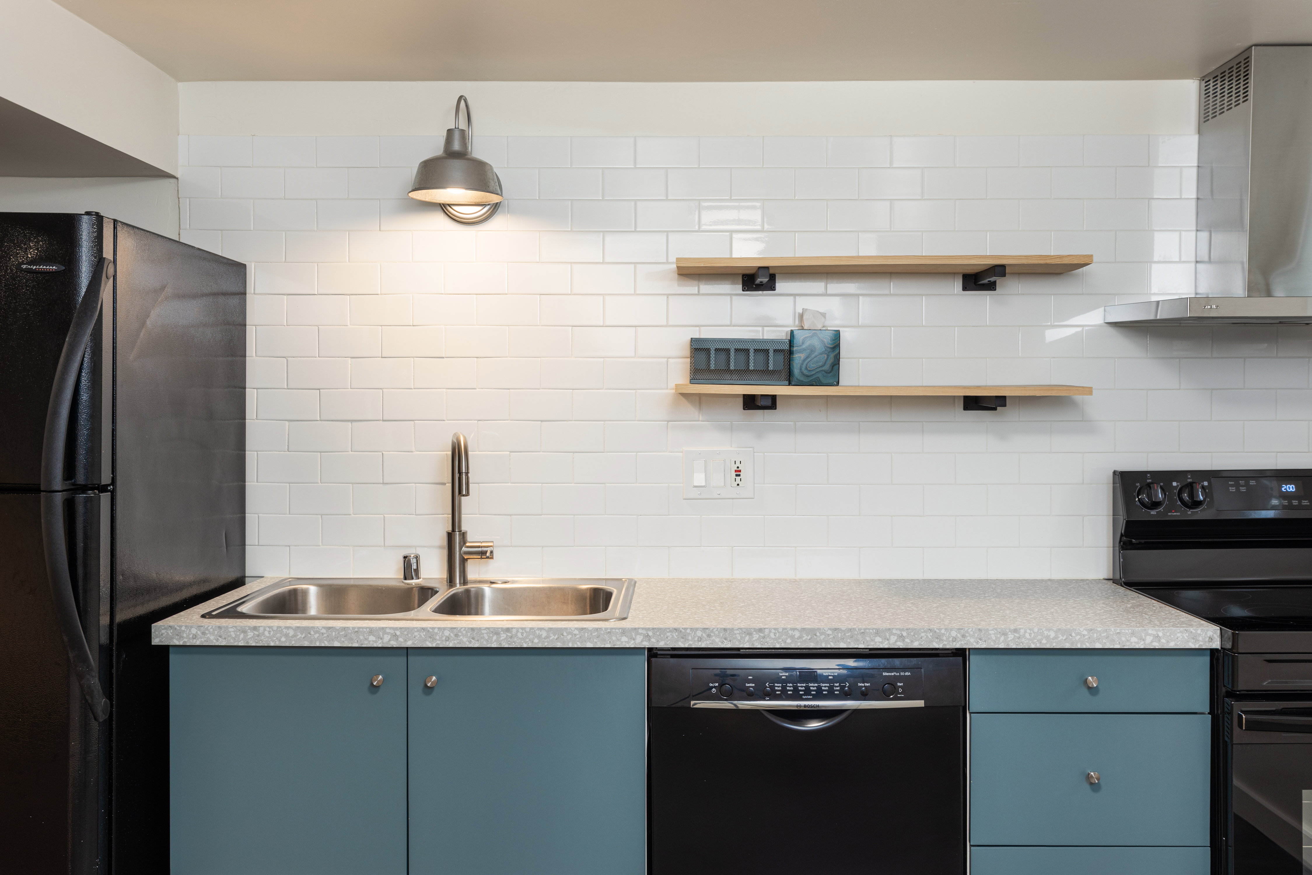 Kitchen featuring a double basin stainless steel sink with a gooseneck faucet, light grey speckled countertops, teal lower cabinetry with silver hardware, a white subway tile backsplash, and two wood-finish floating shelves