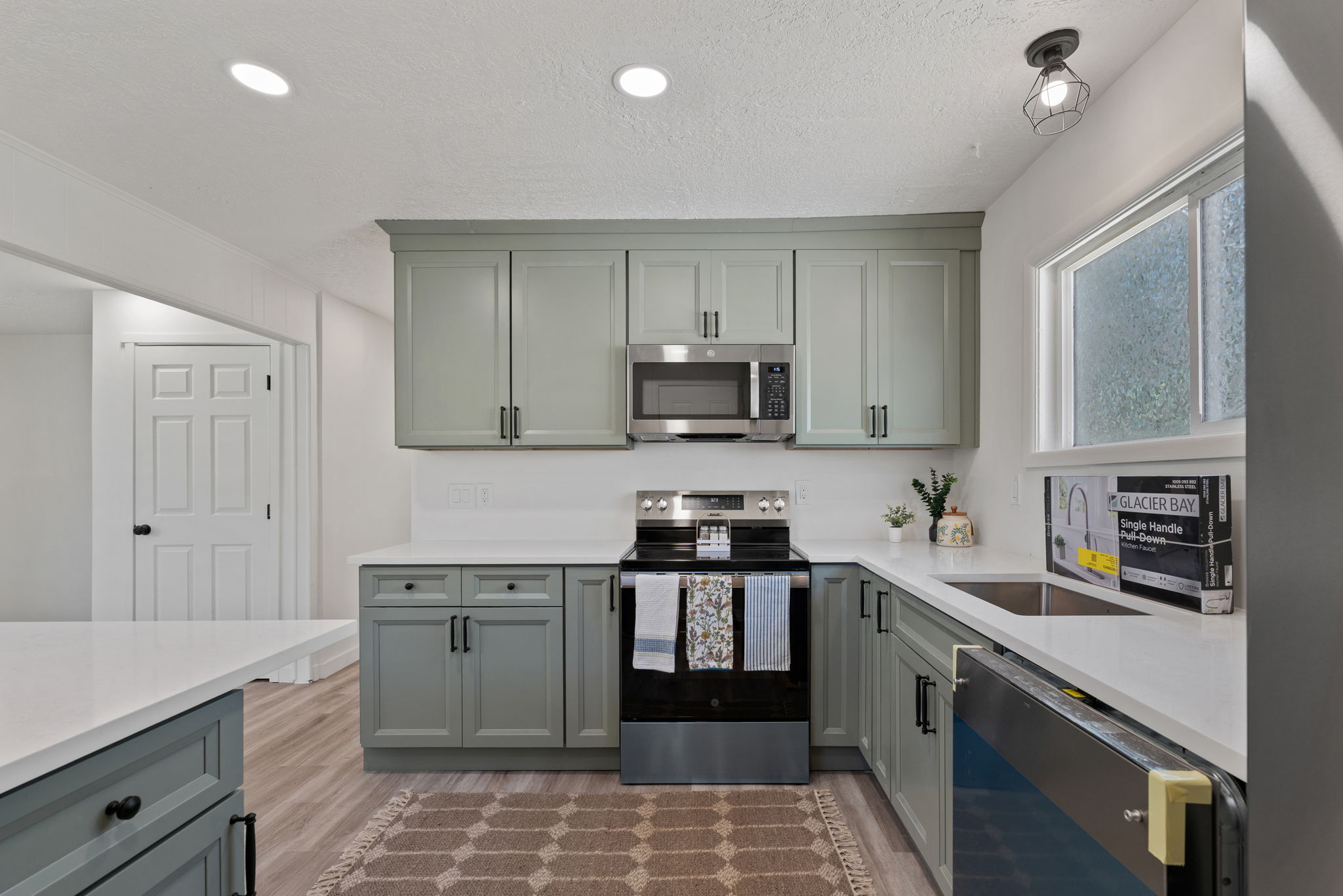Modern kitchen featuring sage green cabinetry, stainless steel appliances including a microwave and range, and light-toned countertops
