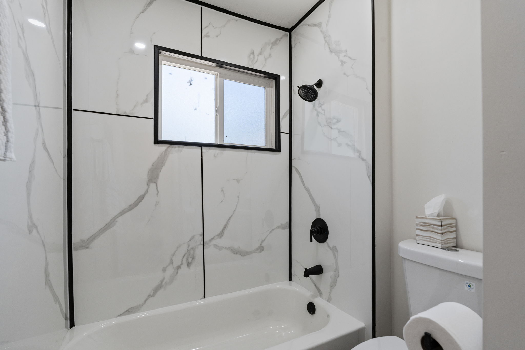 Bathtub and shower combination featuring white marble-look paneling with black trim, a black-framed window, and matte black fixtures