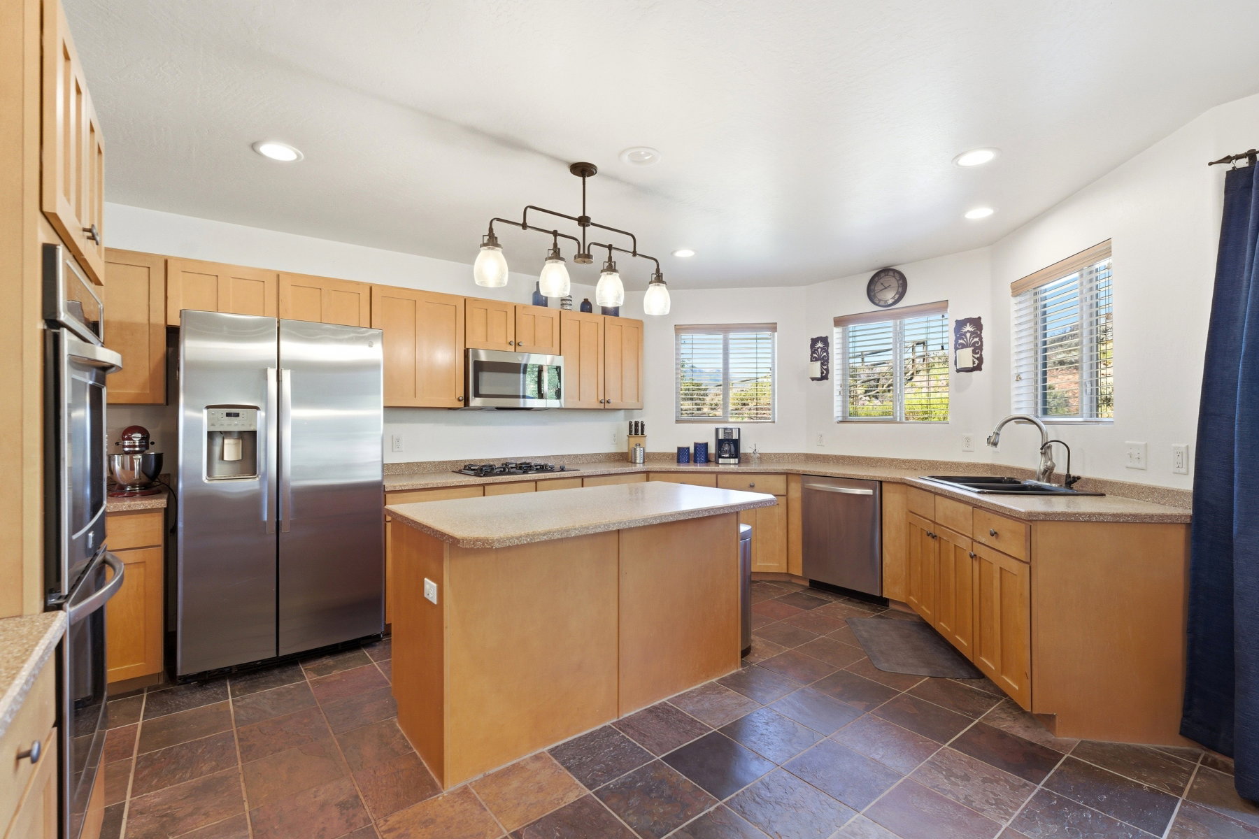 Kitchen featuring a central island, light wood-finish cabinetry, stainless steel appliances, stone-look countertops, and dark tile flooring