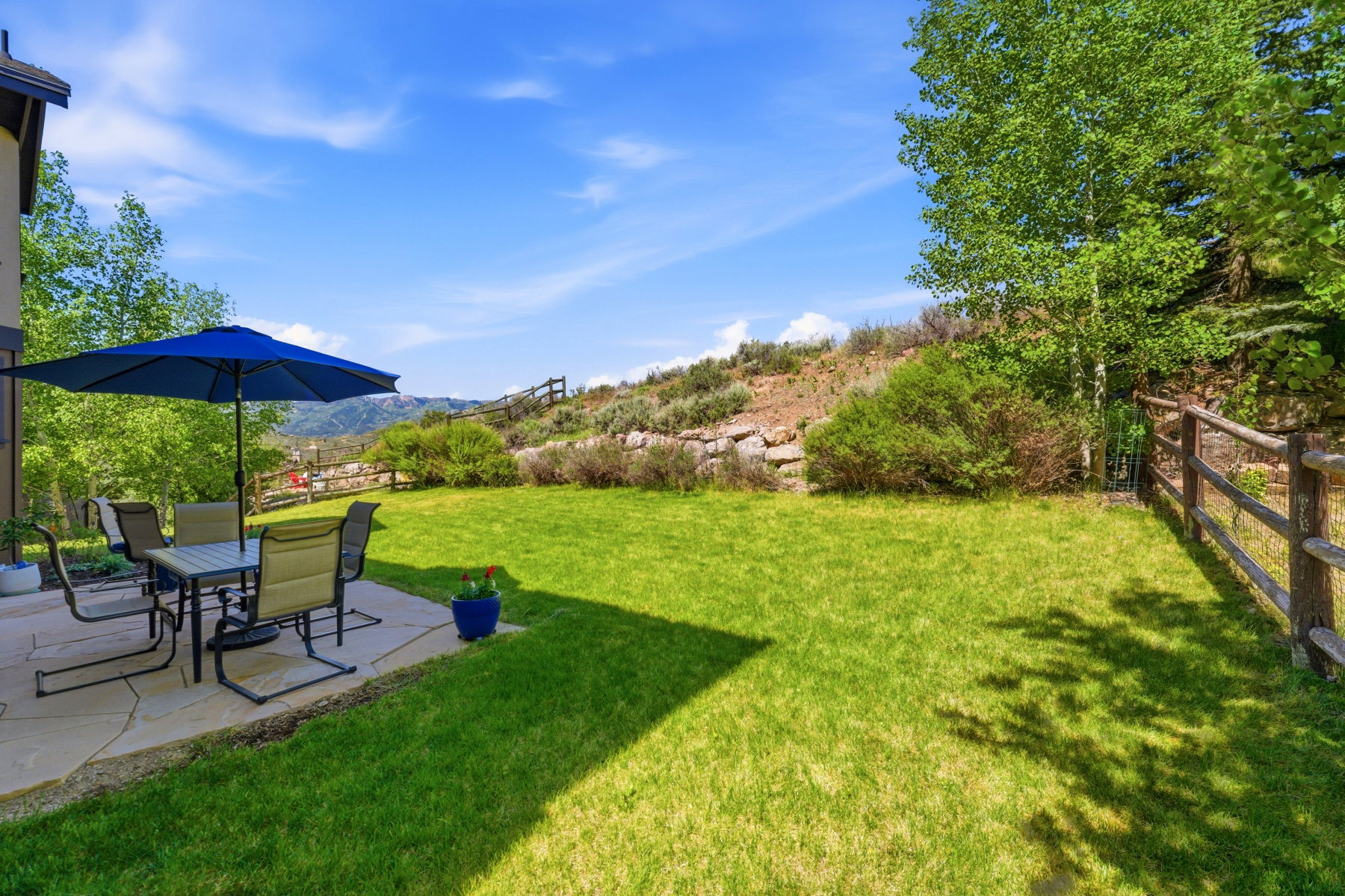Expansive backyard featuring a stone patio, vibrant green lawn, mature trees, and a split-rail fence