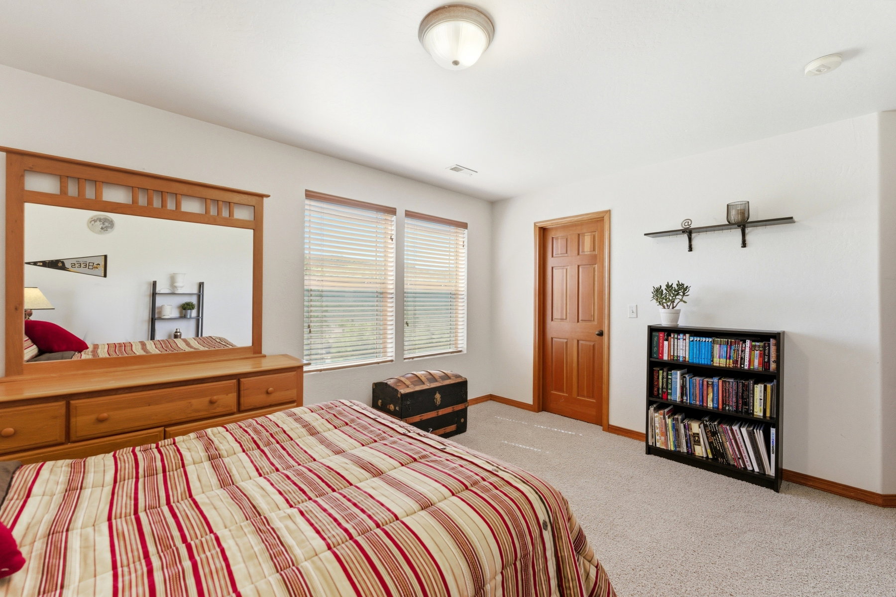 Carpeted room featuring a ceiling-mounted light fixture, two windows with blinds, a wood-paneled door, and a wall-mounted shelf