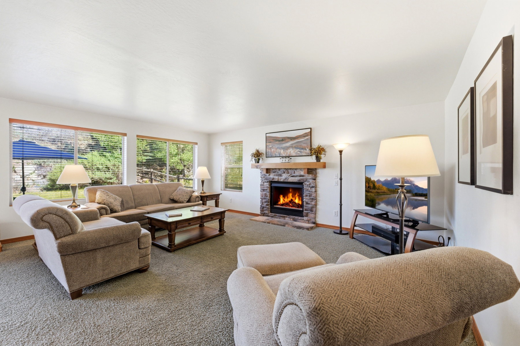 Living area featuring a stone fireplace with a wood mantle, large windows with horizontal blinds, and light-colored carpeting
