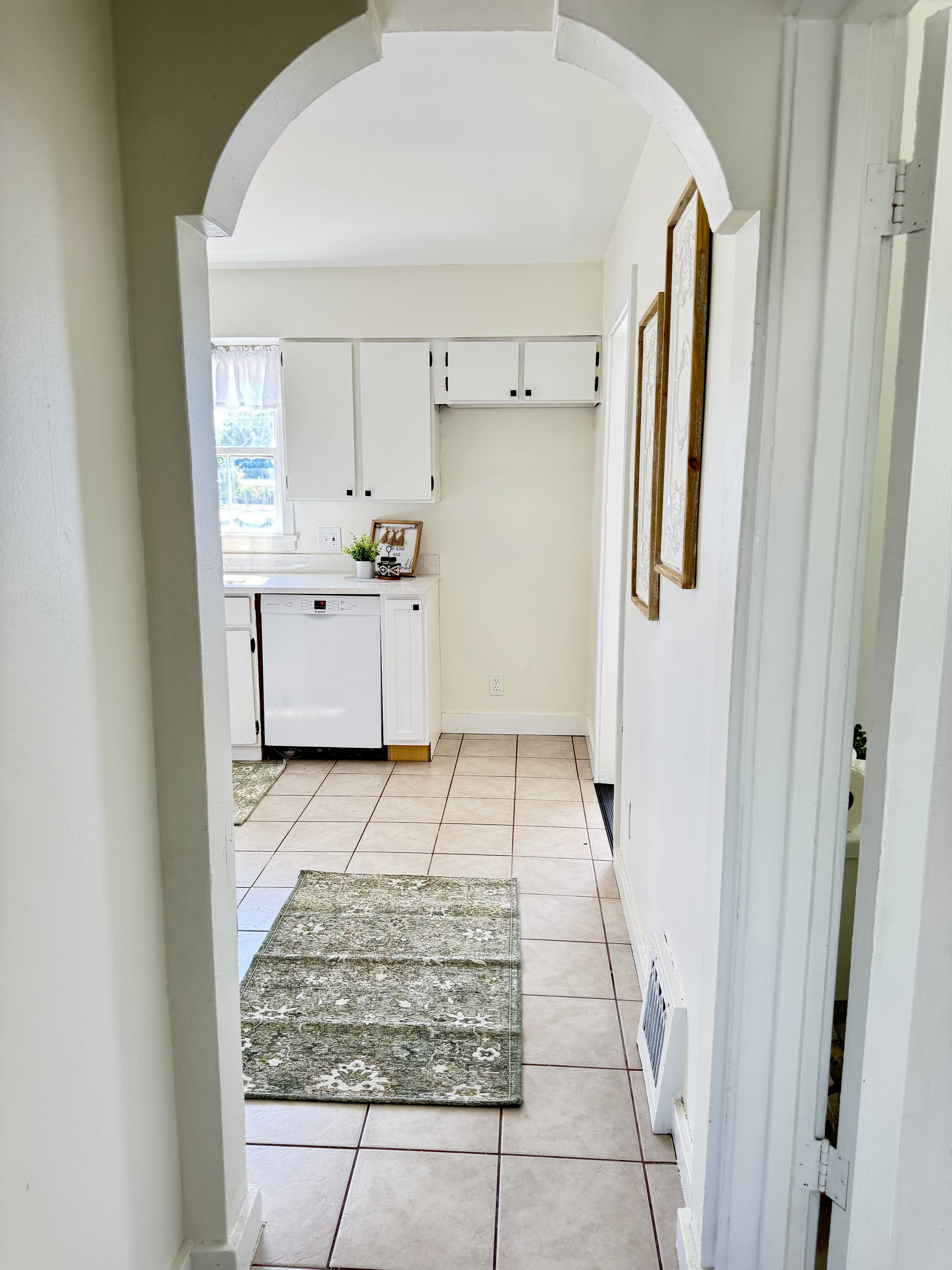 Archway entryway to a kitchen featuring square tile flooring, white cabinetry with black hardware, a white dishwasher, and a window with natural light
