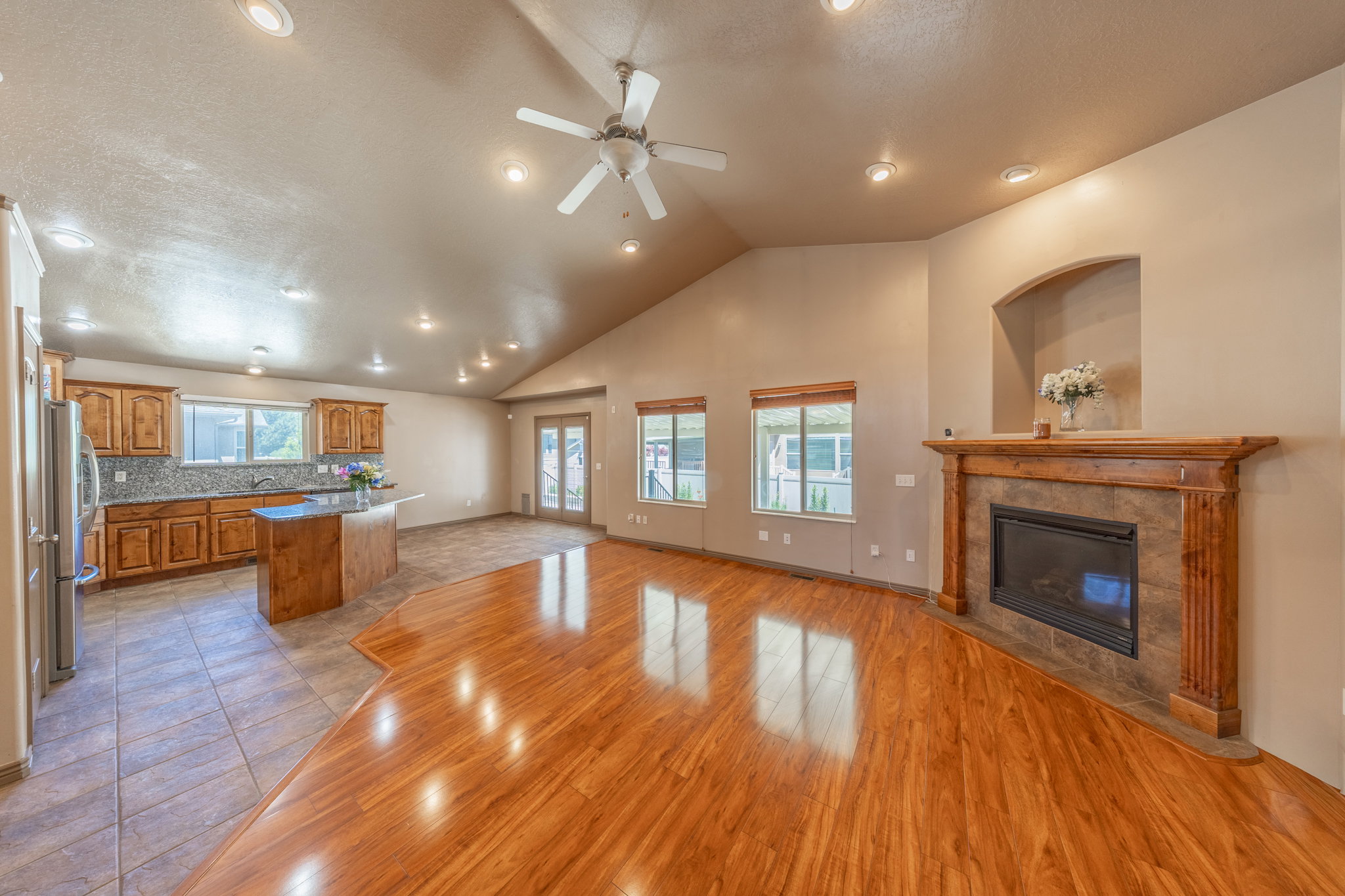Open-concept living area featuring a vaulted ceiling with recessed lighting and a ceiling fan, wood-finish flooring, and a fireplace with a wood mantel and stone surround
