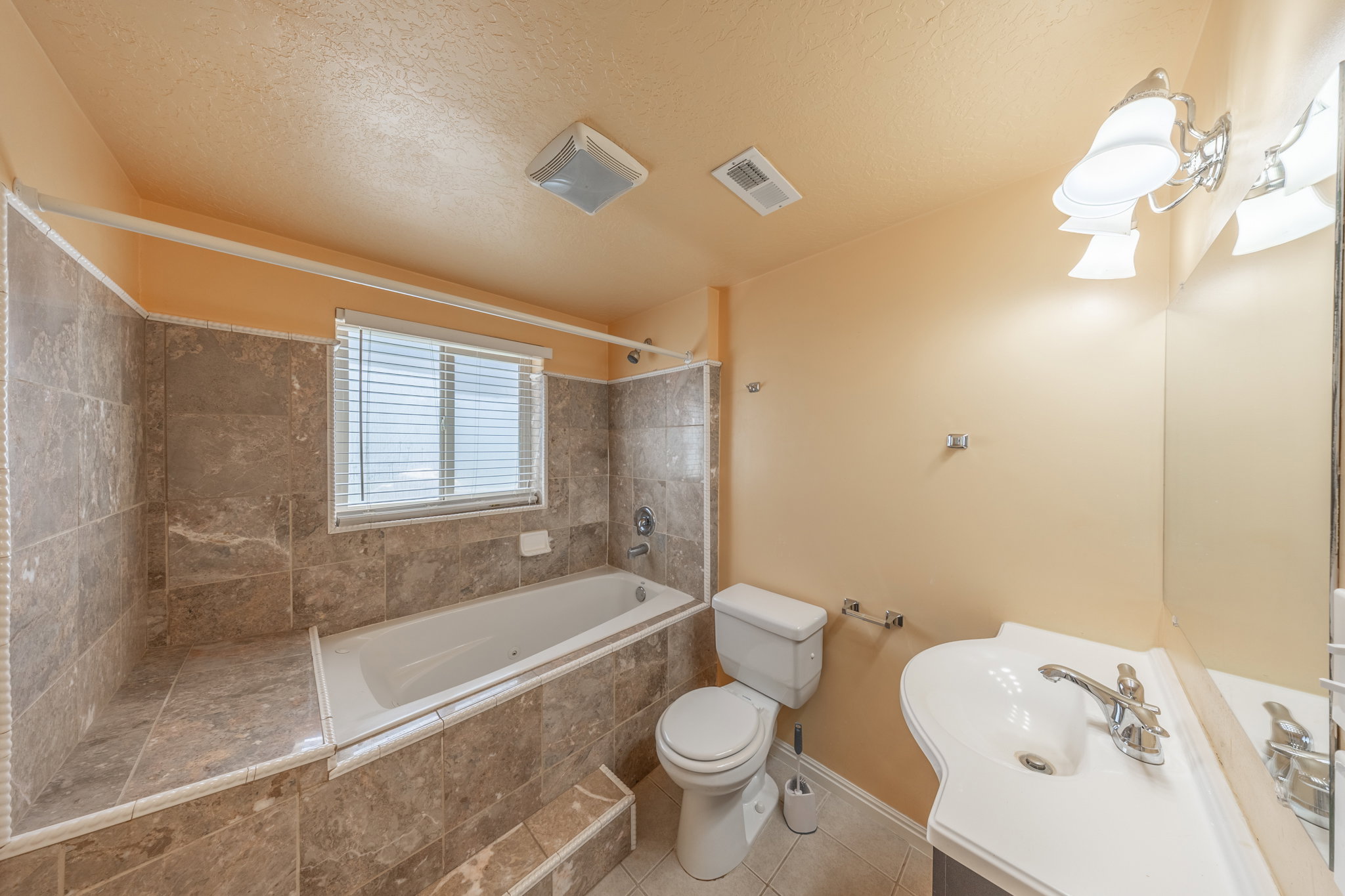 Bathroom featuring a built-in bathtub with tiled surround, a window with blinds, a toilet, and a vanity with an integrated sink and chrome faucet