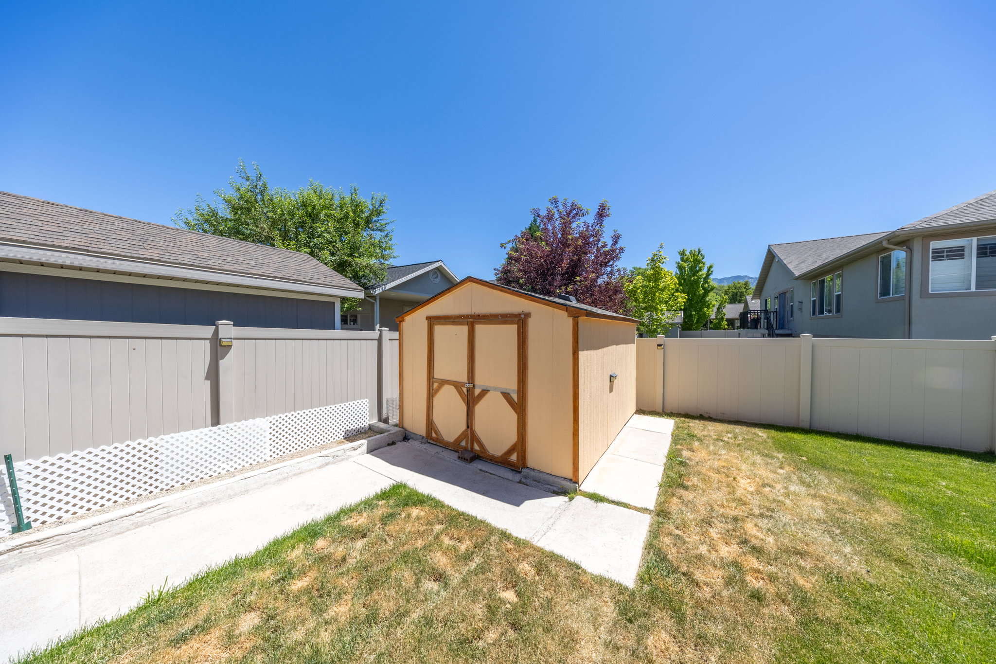 Backyard featuring a light-colored storage shed with dark trim, concrete pathways, and surrounding vinyl fencing
