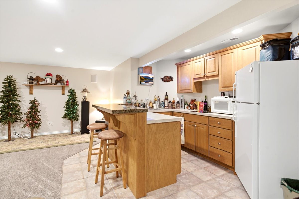 Built-in bar with stone-look countertop and seating, complemented by a full kitchen area featuring wood cabinetry, light-toned countertops, and tile flooring