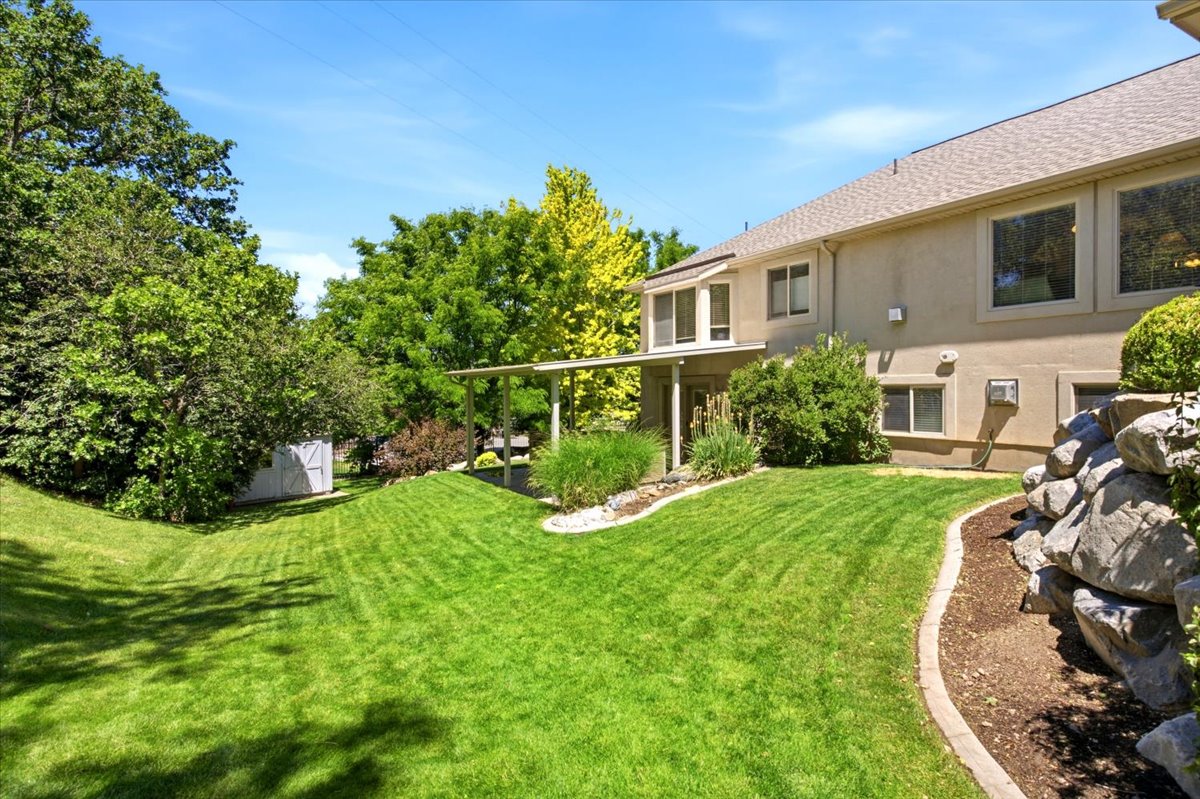 Expansive backyard featuring a lush lawn, mature trees, rock landscaping, a covered patio area, and a detached storage shed