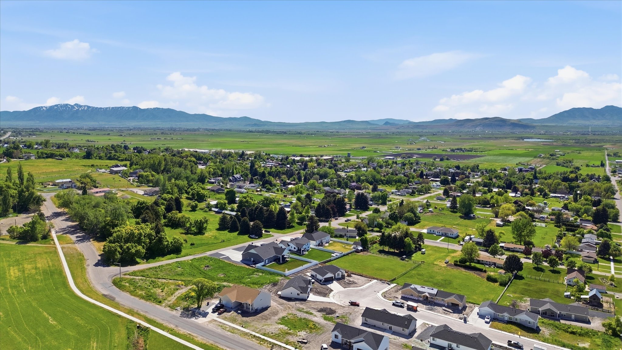 Aerial depiction of a developing community with new construction homes, established residences, expansive green fields, mature trees, and distant mountains