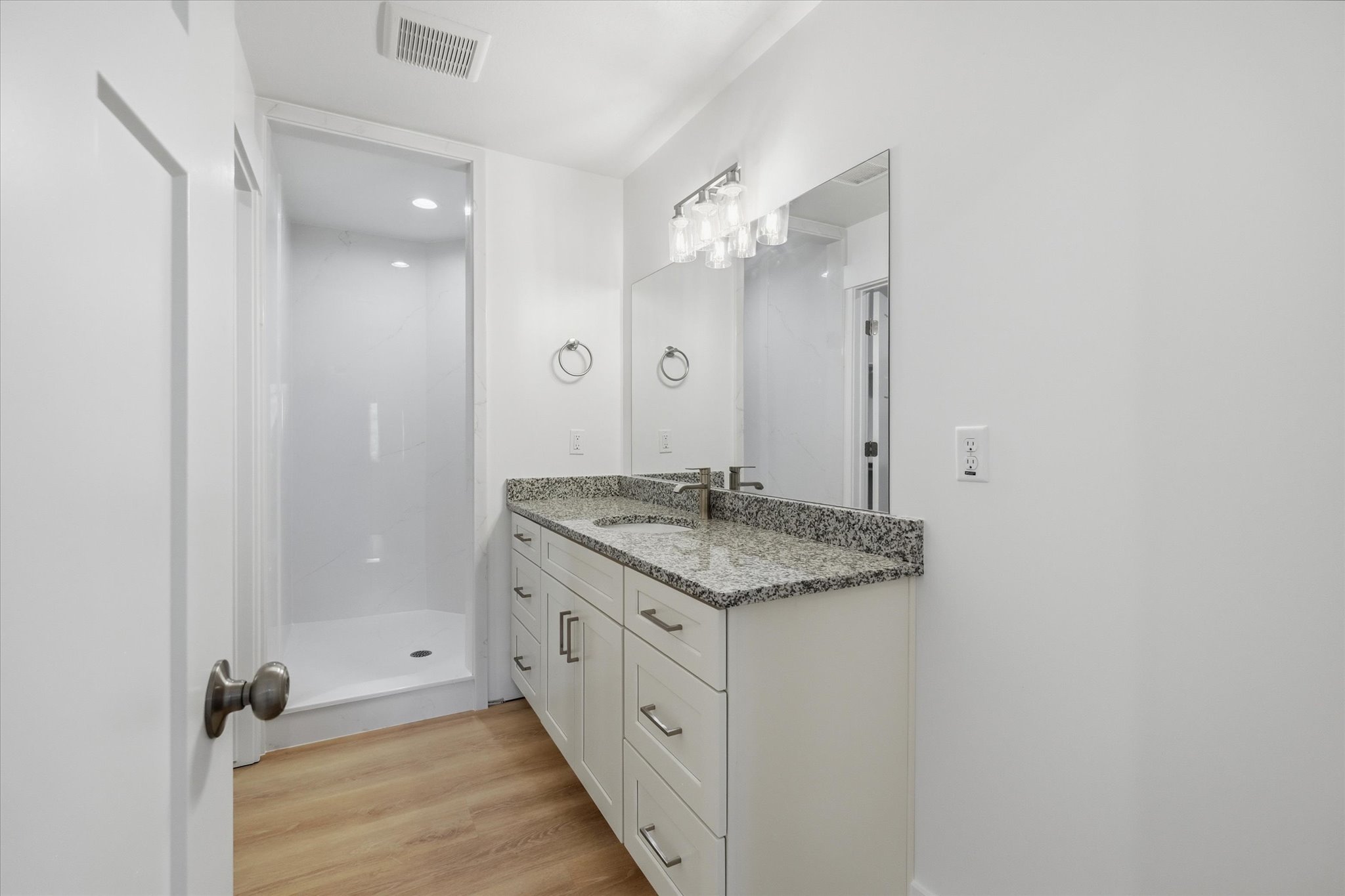 Bathroom featuring a walk-in shower with white surround, a vanity with a speckled countertop and integrated sink, a rectangular mirror, and wood-finish flooring